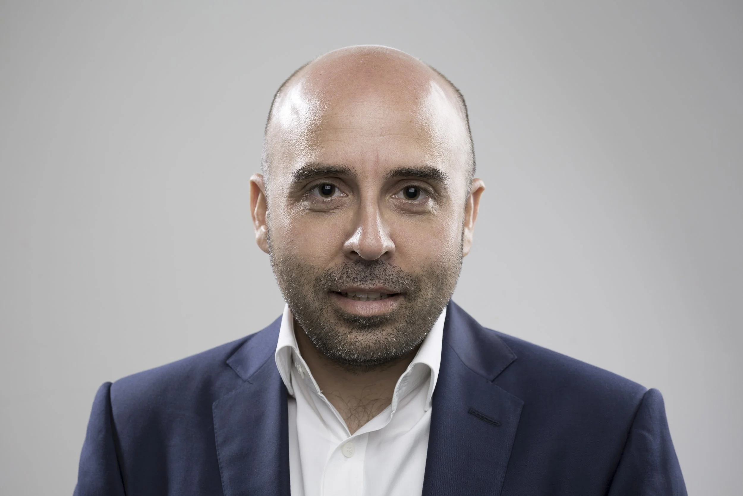 Headshot of a bald man with light beard, wearing a white shirt and navy blazer, against a plain grey background.