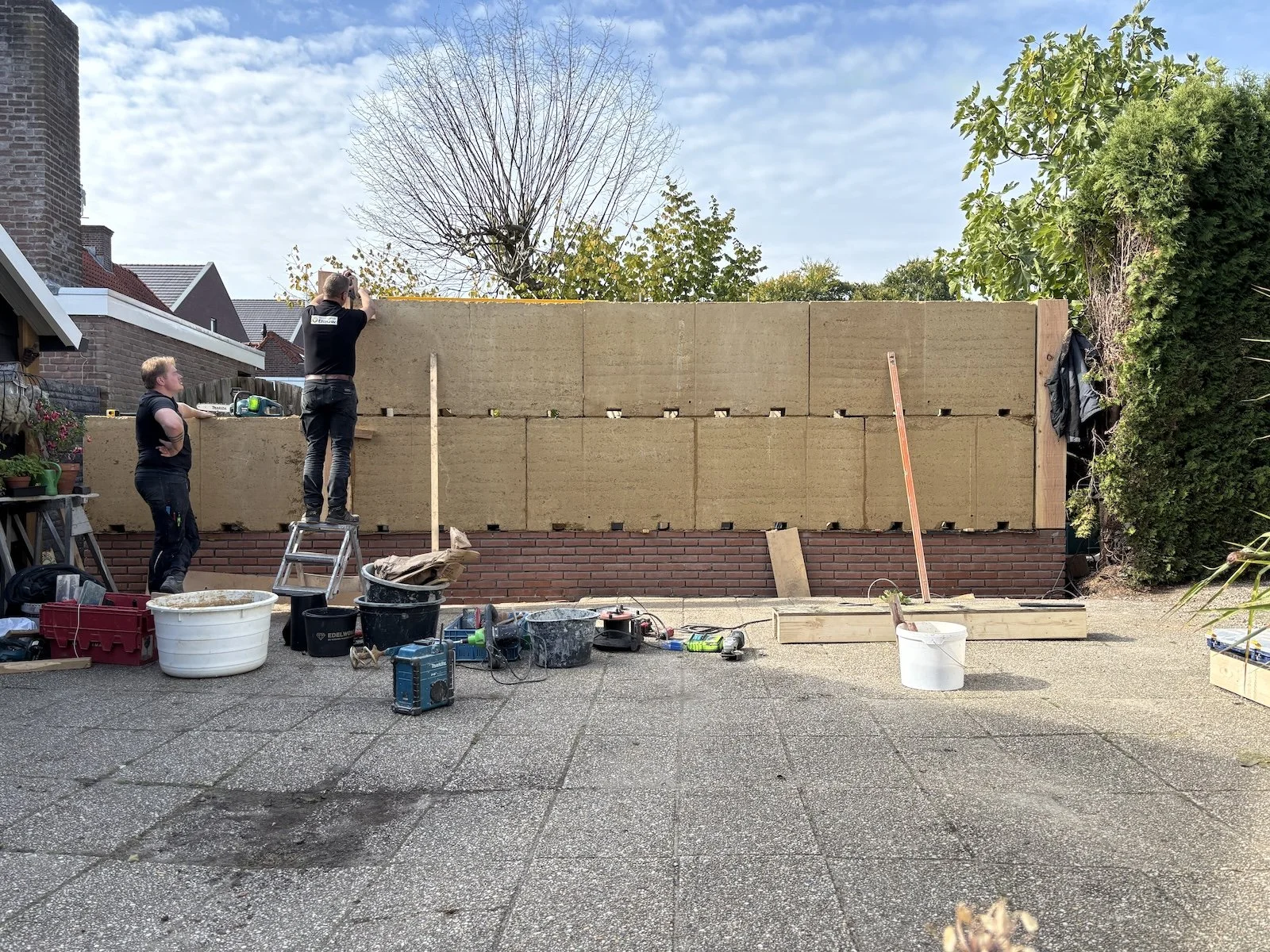 Twee mensen bouwen een muur uit stenen en isolatiemateriaal op een terras. Er staan verschillende bouwgereedschappen en materialen op de grond.