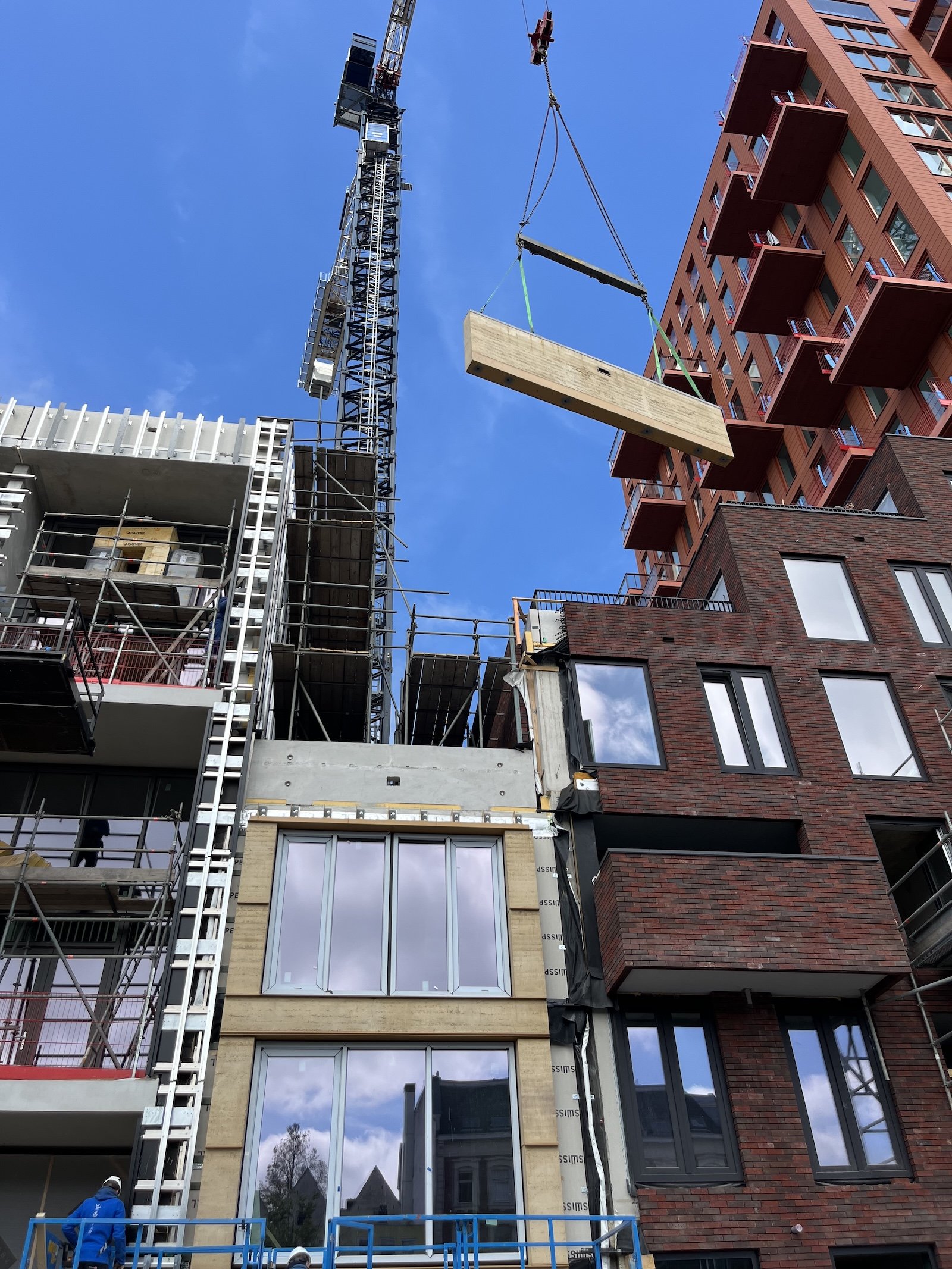 Een hoogbouwproject met een bouwkraan die een groot betonelement optilt, omgeven door steigers en een nieuw gebouw met glazen ramen en rode bakstenen gevels.