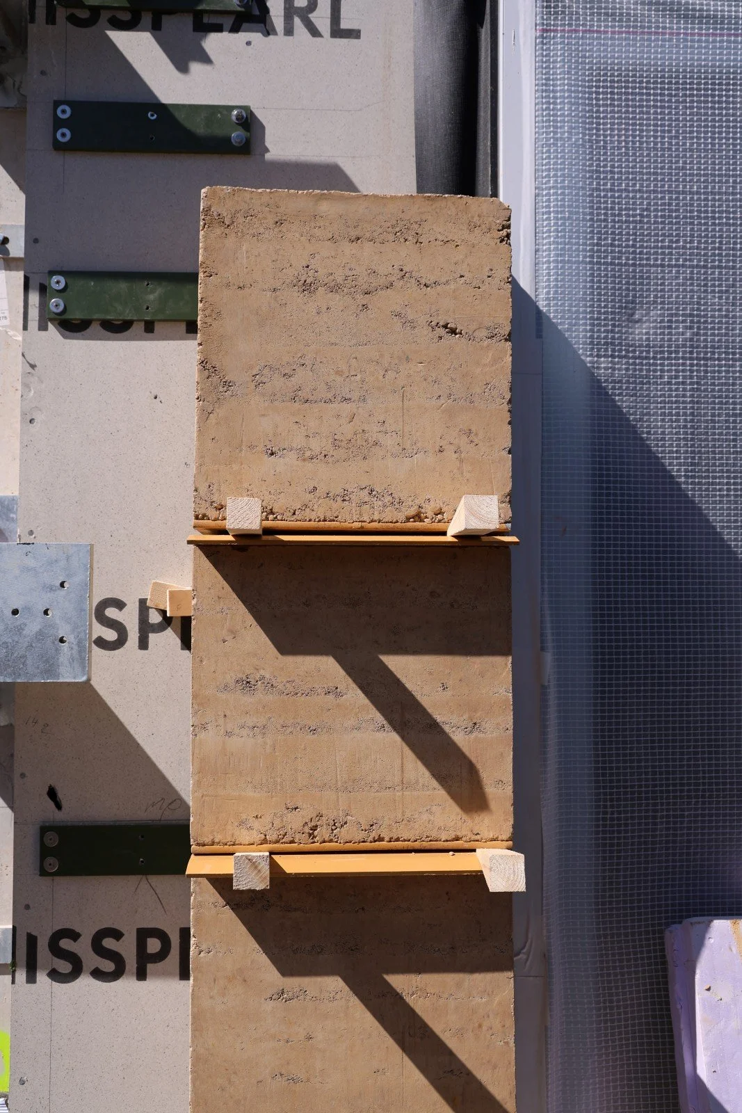 Een foto van verschillende bouwmaterialen, waaronder twee grote stenen tegels, een stuk isolatieplastiek, metalen bevestigingsstukken en een stuk gaas. De tegels worden ondersteund door kleine houten blokken.