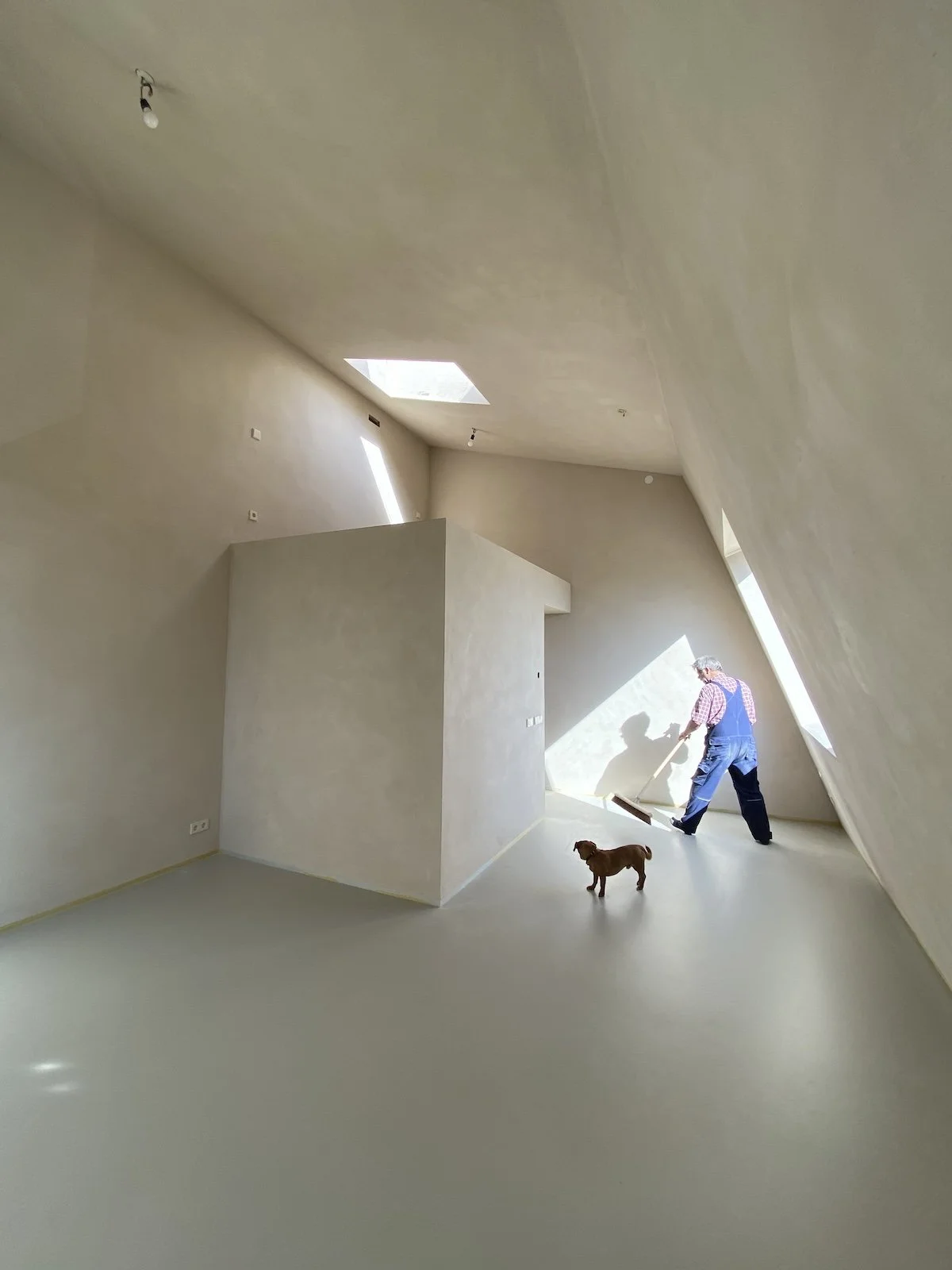 Een man die de vloer veegt in een kamer met schuine plafonds en grote ramen, met een hond erbij.