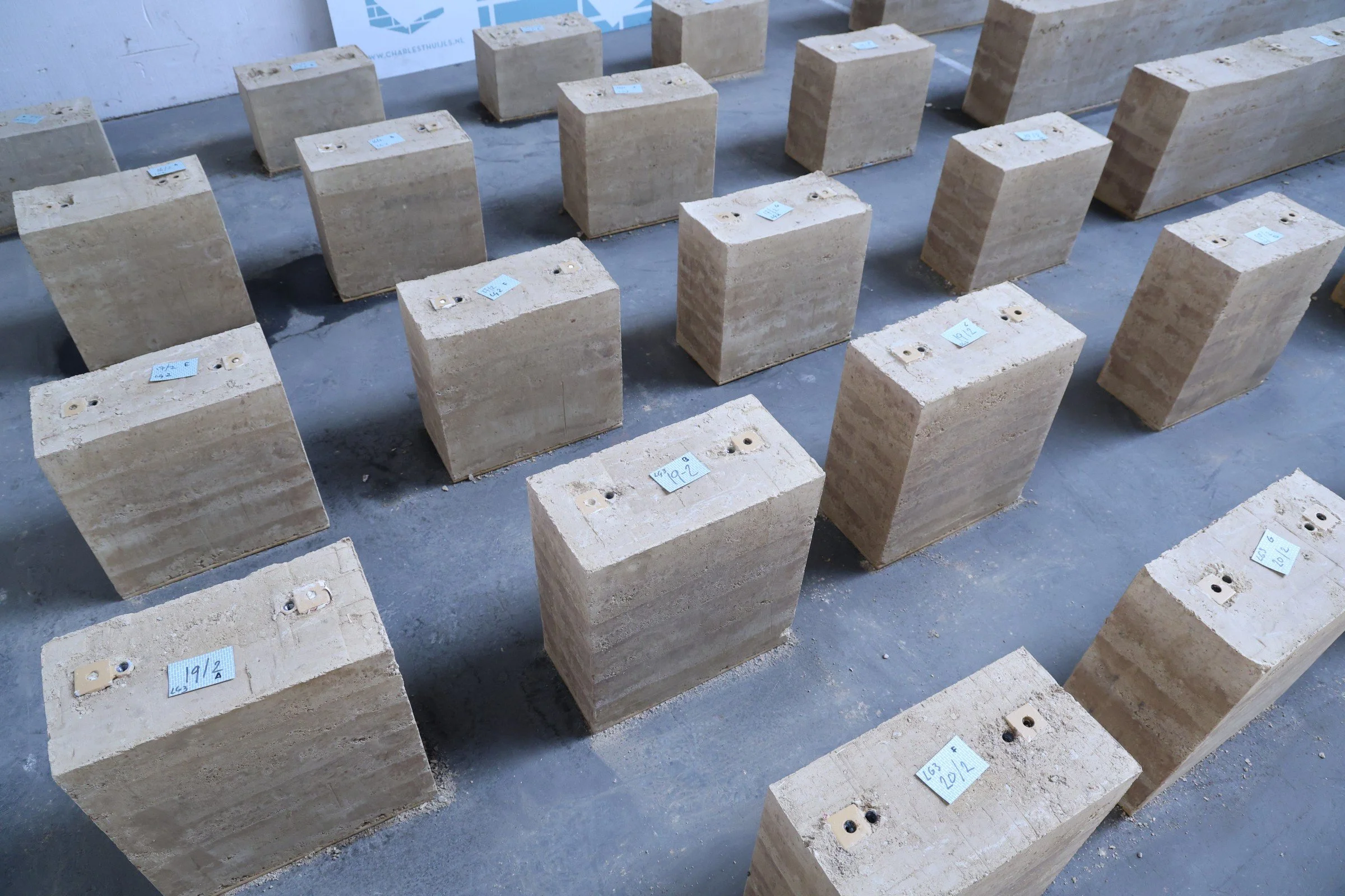 Rijen grote zandstenen blokken met indicatielabels en eventuele bevestigingspunten, geplaatst op een betonvloer in een bouw- of restauratieomgeving.
