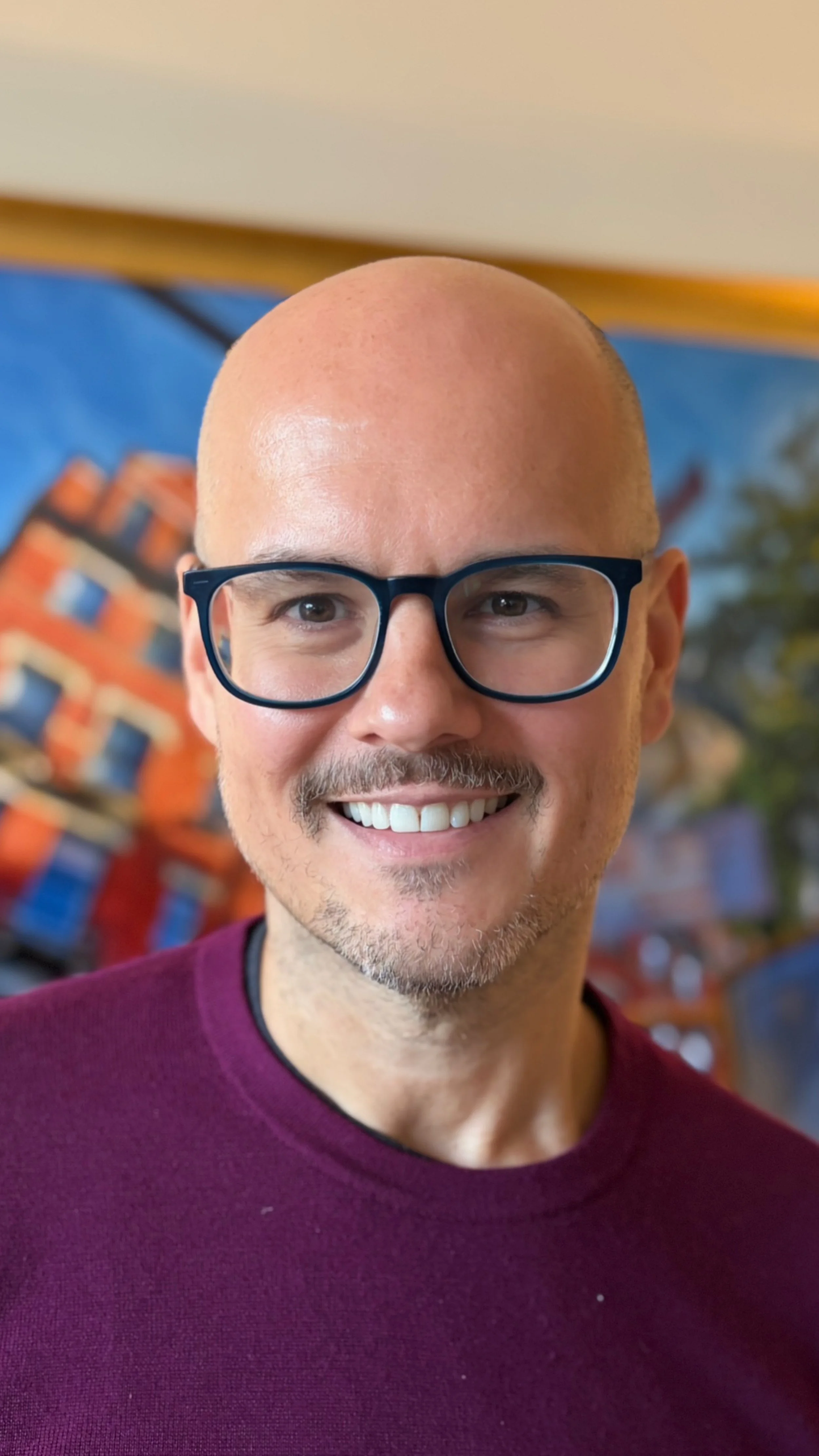 A smiling man wearing a maroon sweater, standing in front of a colorful mural.
