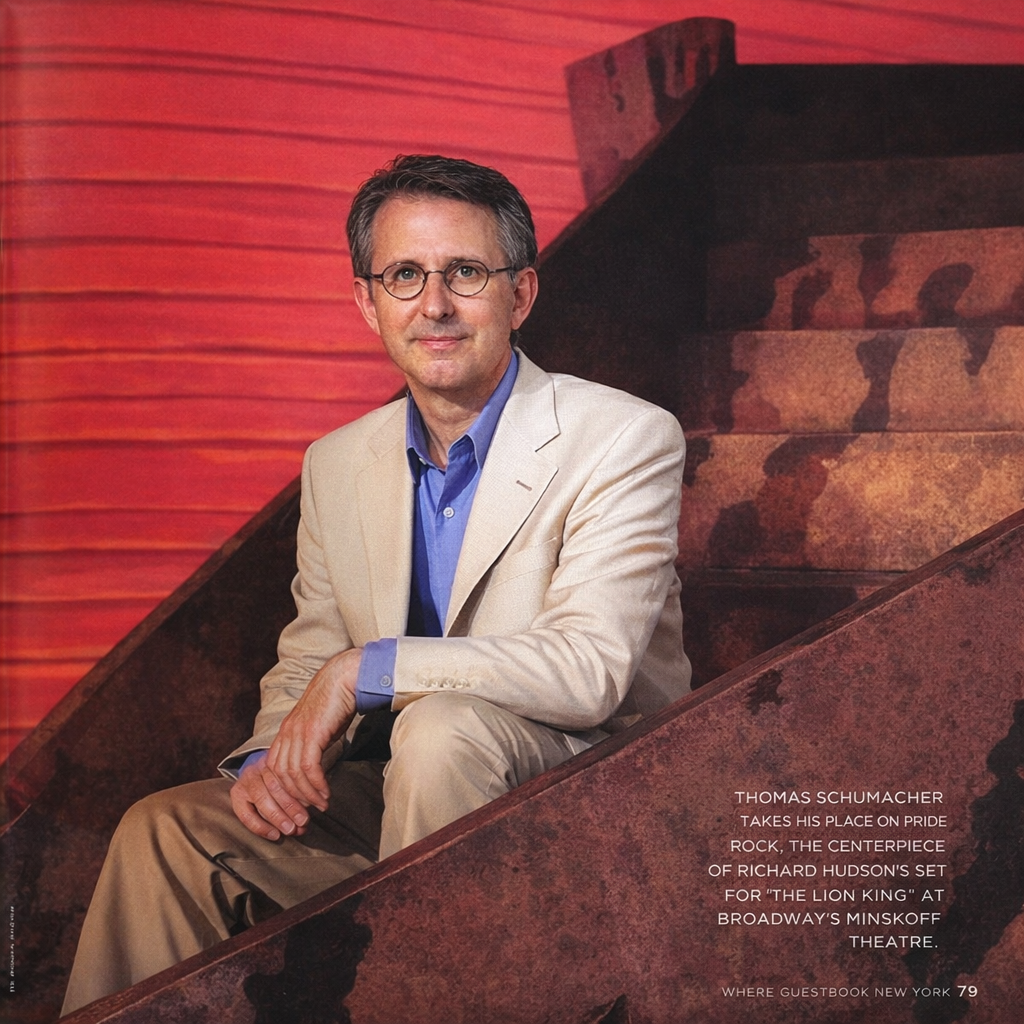 A man in a beige suit and blue shirt sitting on theater stairs with a red background. Text indicates it is Thomas Schumacher at Broadway's Minskoff Theatre for "The Lion King."