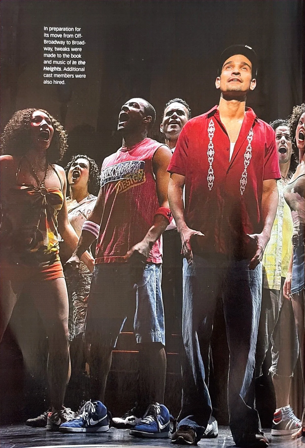 Group of diverse actors on stage during rehearsal for a musical, with some singing or speaking, and some looking upward, wearing casual clothing, with the background as a black curtain.
