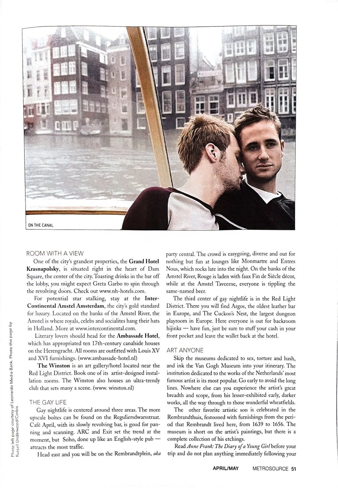 Two young men sitting closely on a boat, with one leaning his head on the other’s shoulder, against the backdrop of a city canal with tall buildings.