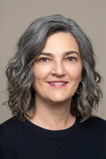 A woman with shoulder-length gray hair, wearing a black top and hoop earrings, smiling at the camera against a plain background.