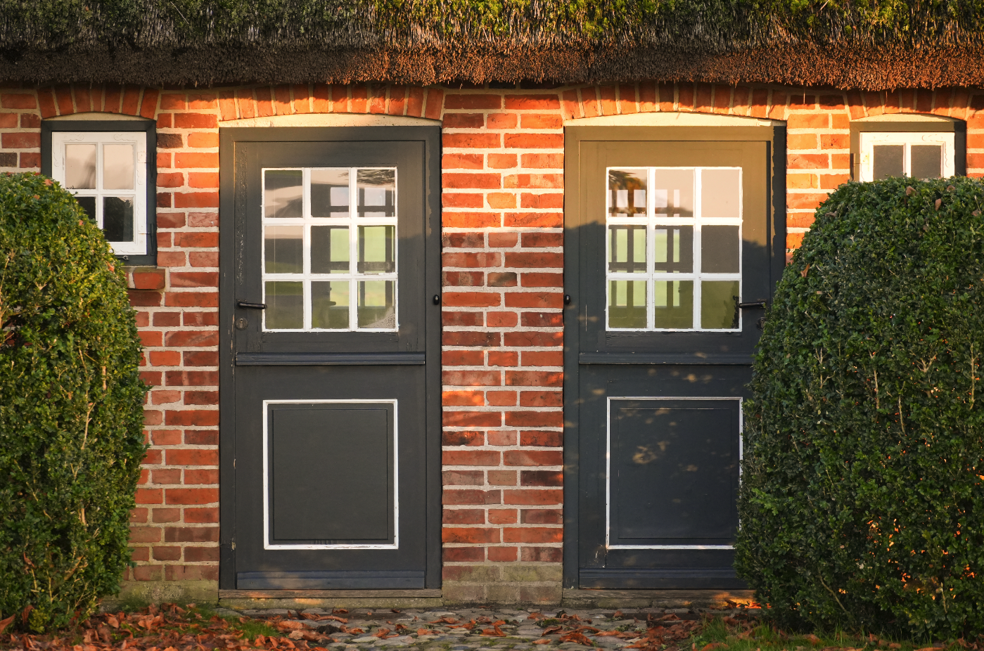 Zwei dunkle Holztüren vor einer roten Backsteinwand, flankiert von grünbuschigen Büschen, mit weißen Fensterläden und einem Strohdach.