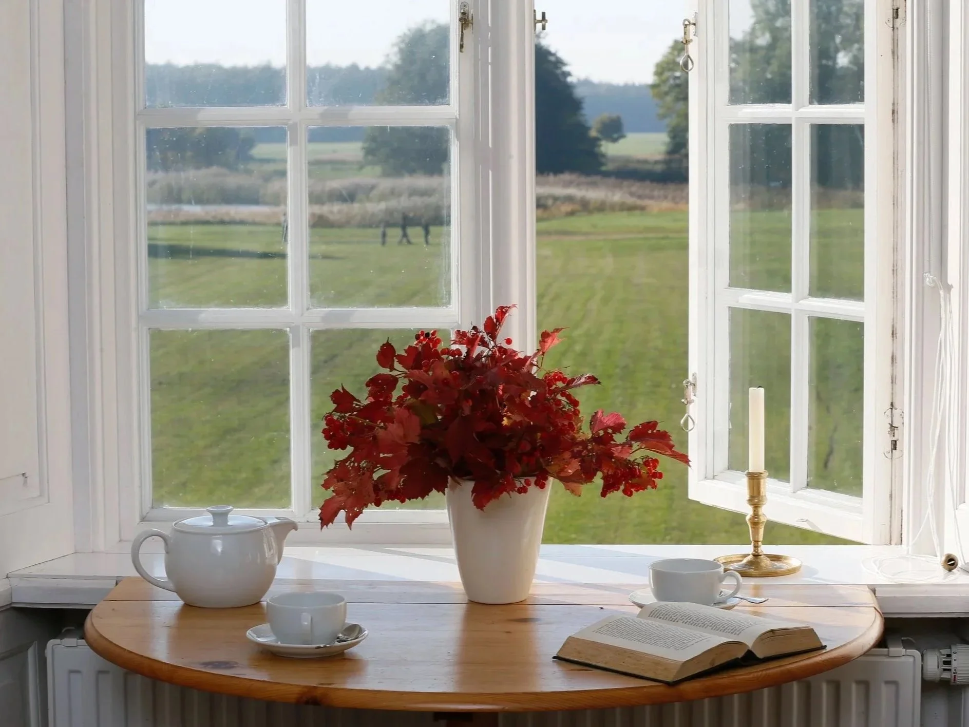 Ein Fenster mit weißen Rahmen, das nach außen geöffnet ist. Auf einem runden Holztisch im Vordergrund stehen eine weiße Kanne, eine Vase mit roten Herbstblättern, eine Tasse und ein offenes Buch, neben einem weiteren weißen Teegeschirr. Im Hintergrun