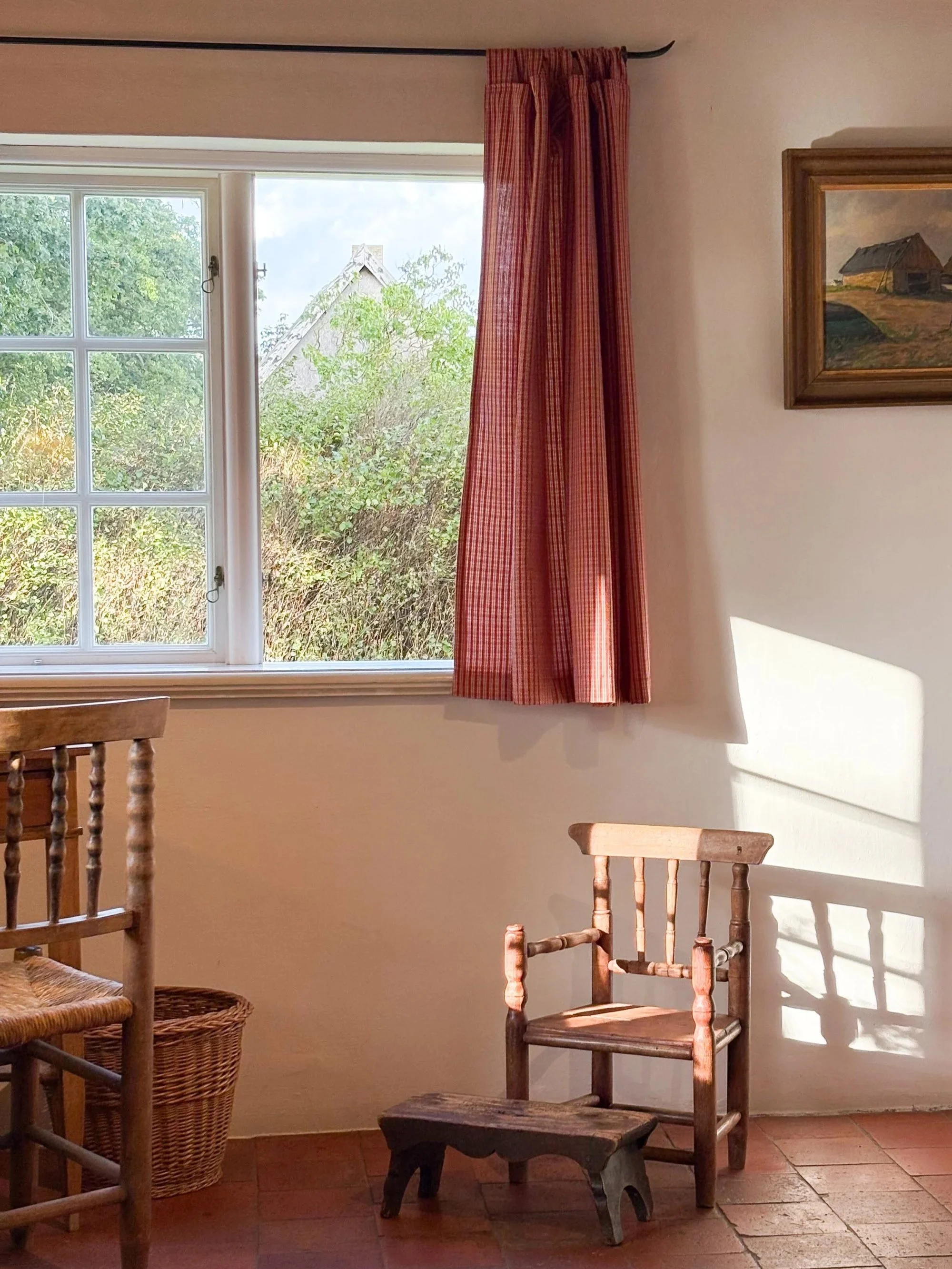 Ein Raum mit einem Fenster, rotem Gardinen, einem Wandbild von Scheunen und Booten, Holzmöbeln, darunter Stuhl und Hocker, auf Terrakottaboden, mit Sonnenlicht, das Schatten wirft.