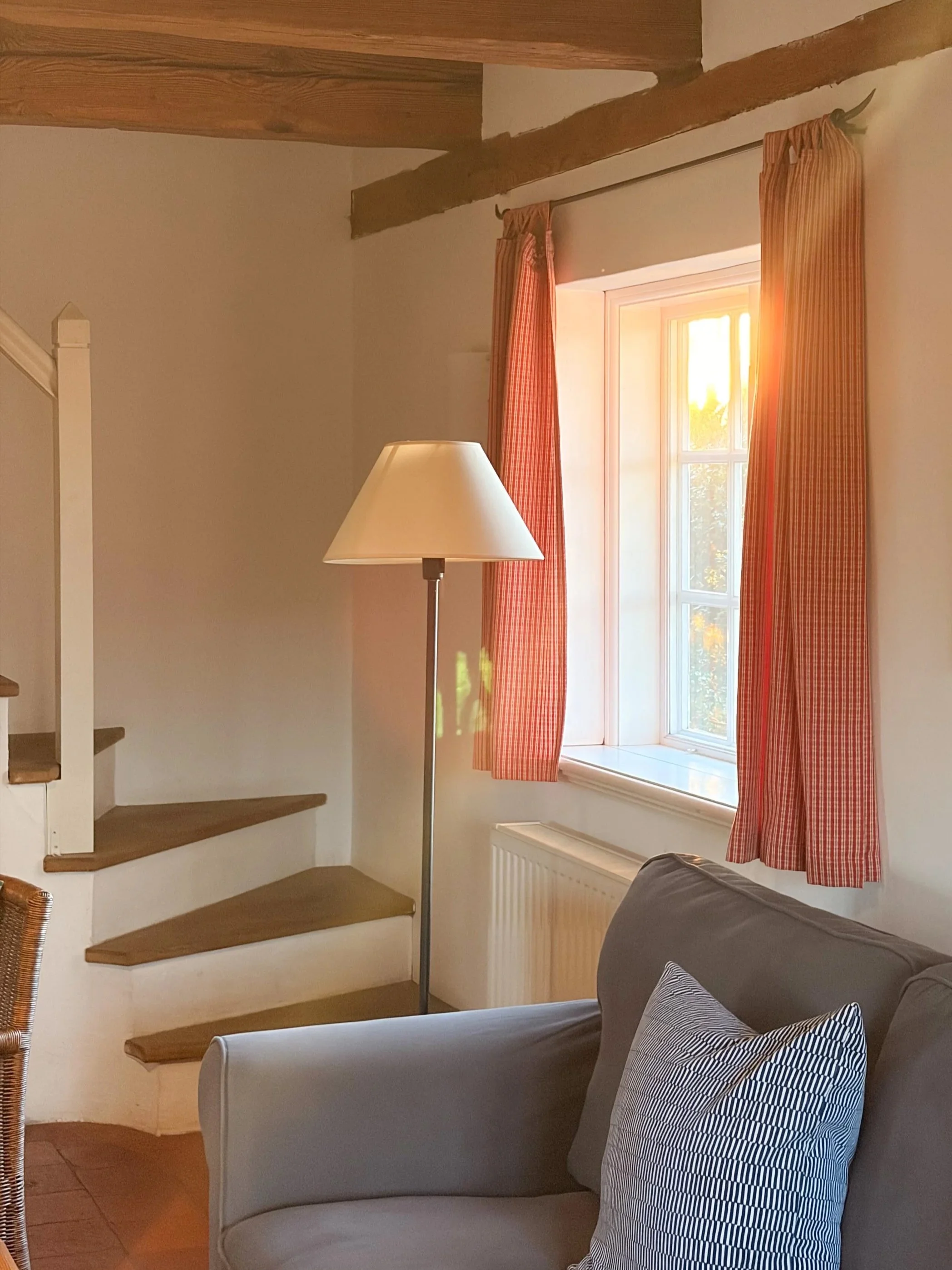 Innenraum mit einem grauen Sofa, einem Kissen mit weiß-blauem Streifen, einem Stehlampen und einem Fenster mit roten und orangefarbenen Gardinen, die Sonnenlicht hereinlassen.