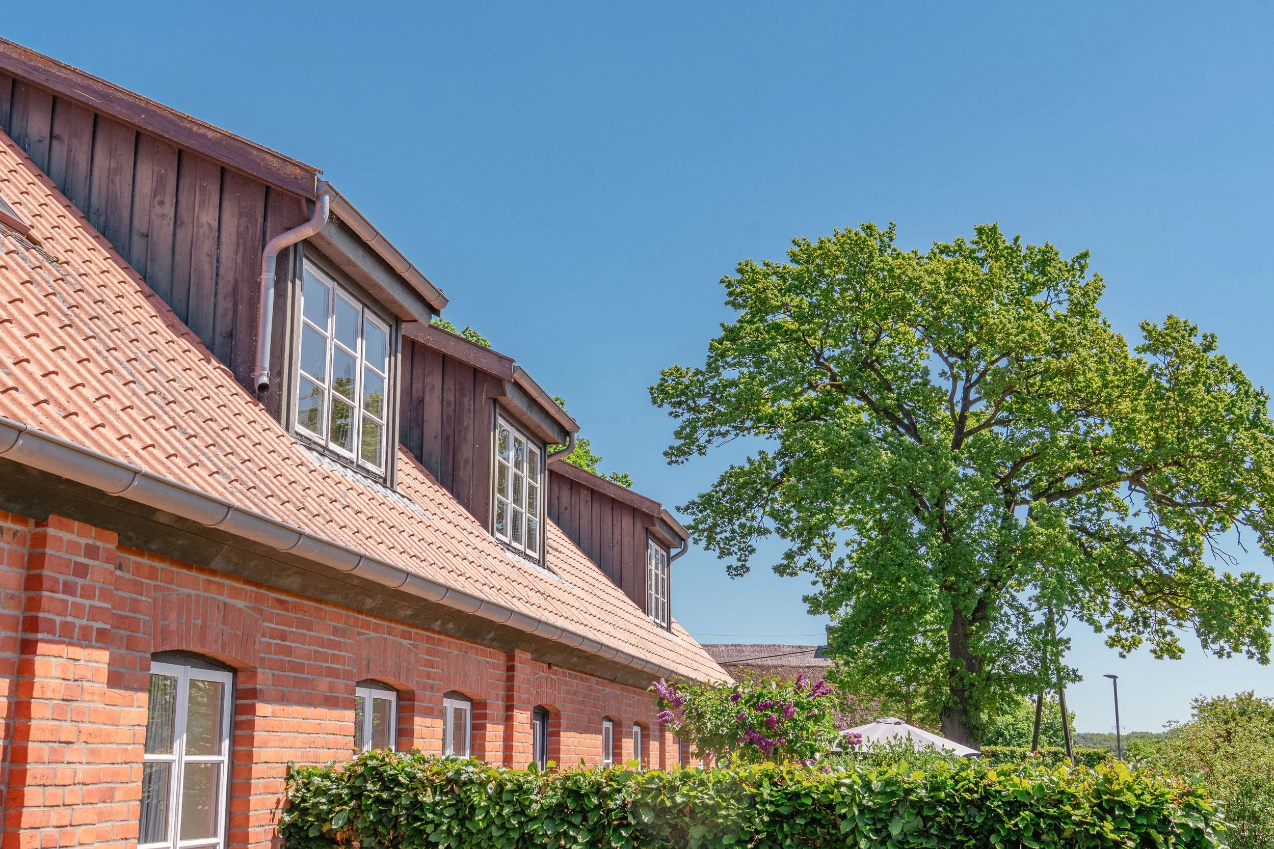 Ziegelhaus mit Gaube unter blauem Himmel, umgeben von grünen Bäumen und Büschen.