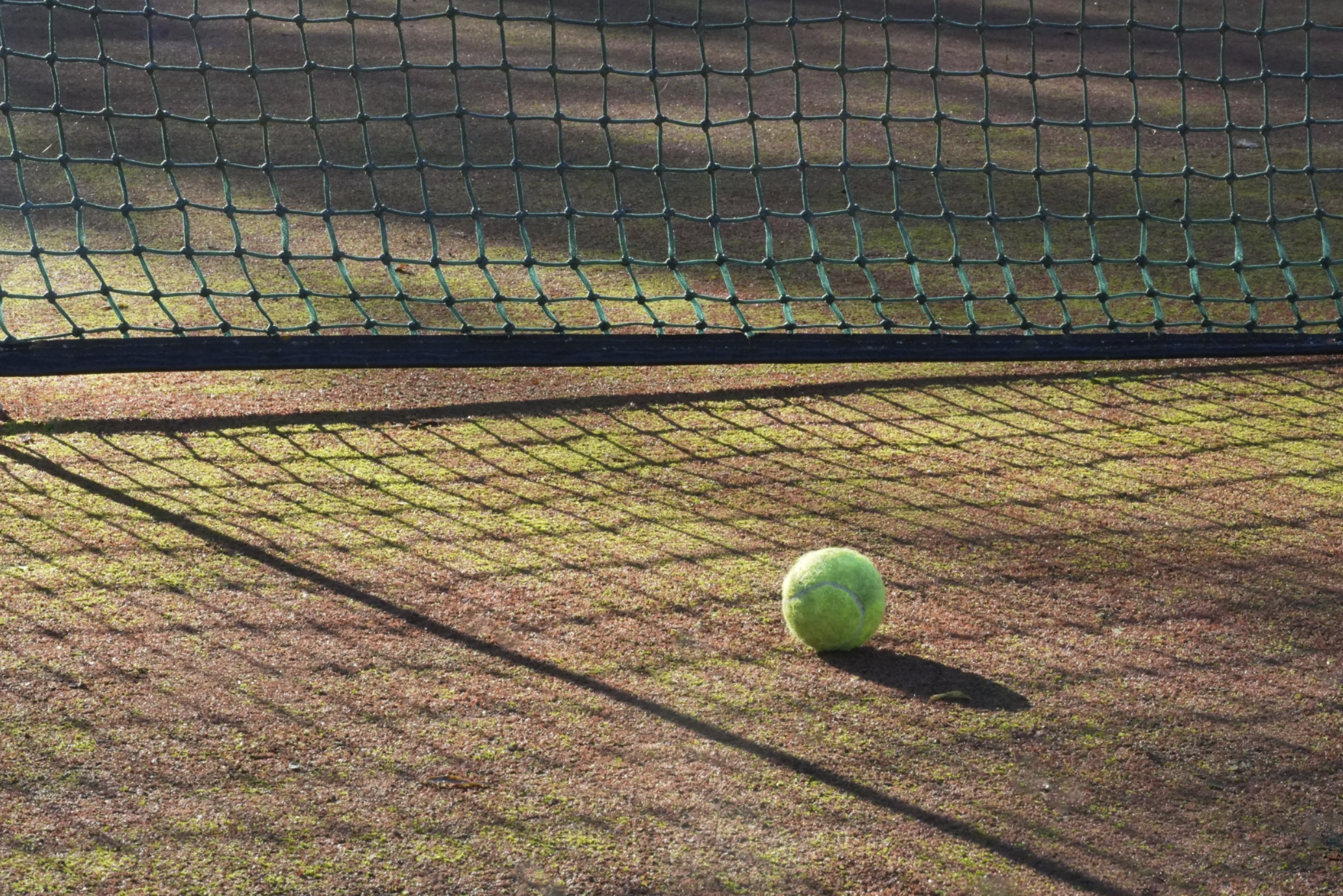 Ein Tennisball liegt auf einem Tennisplatz vor einem Tennisnetz, das im Schatten liegt. Das Netz wirft Schatten auf den Platz.