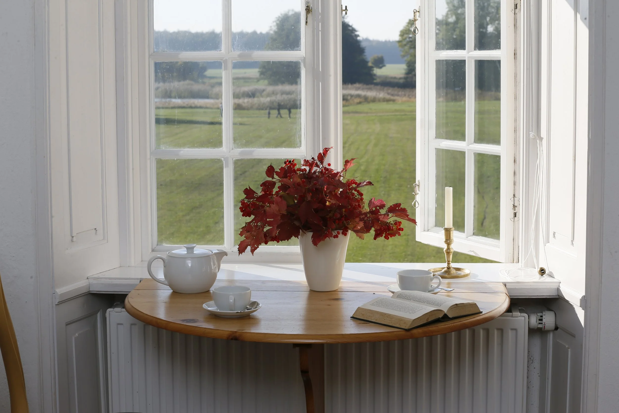 Blick aus einem offenen Küchenfenster auf eine grüne Landschaft mit Bäumen und Wiesen. Auf dem Fensterbrett stehen eine weiße Kanne, eine Vase mit roten Herbstblättern, ein Kerzenhalter mit einer weißen Kerze, ein geöffnetes Buch und eine Tasse.