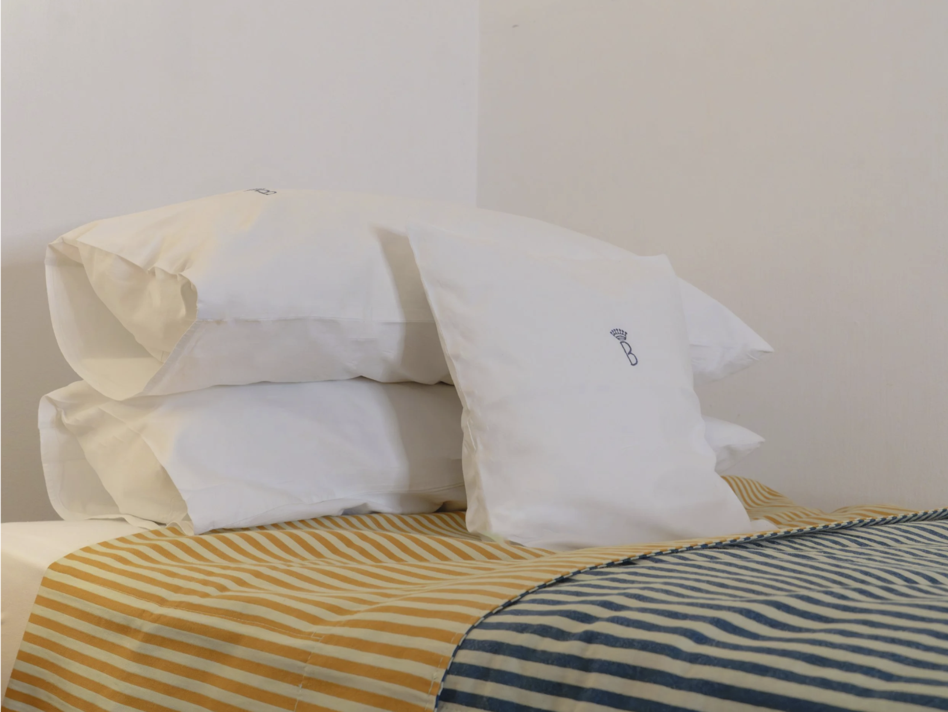 Blick auf ein Bett mit mehreren weiße aufgefaltete Bettwäsche, die teilweise an einem weißen Bettgestell liegen. Das Bett befindet sich an einer weißen Wand. Unterhalb des Betts liegt eine Decke oder ein Bettbezug mit orange-weißen Streifen, der den Fußbereich des Betts bedeckt.