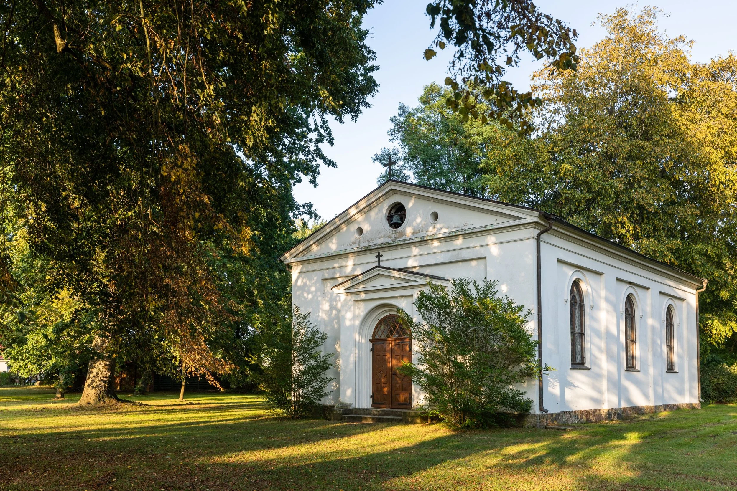 Eine weiße Kapelle auf dem Land in Boldevitz mit hohen Fenstern und Eingangstür, umgeben von grünen Bäumen und einer Wiese im Sonnenschein.