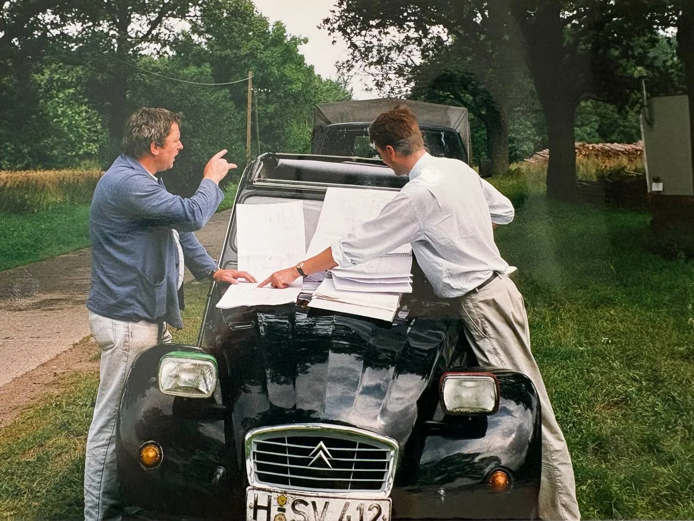 Zwei Männer diskutieren vor einem schwarzen Auto, das auf einer Straße mit Bäumen im Hintergrund parkt. Das Auto ist voller Papierstapel auf dem Motorhaube.