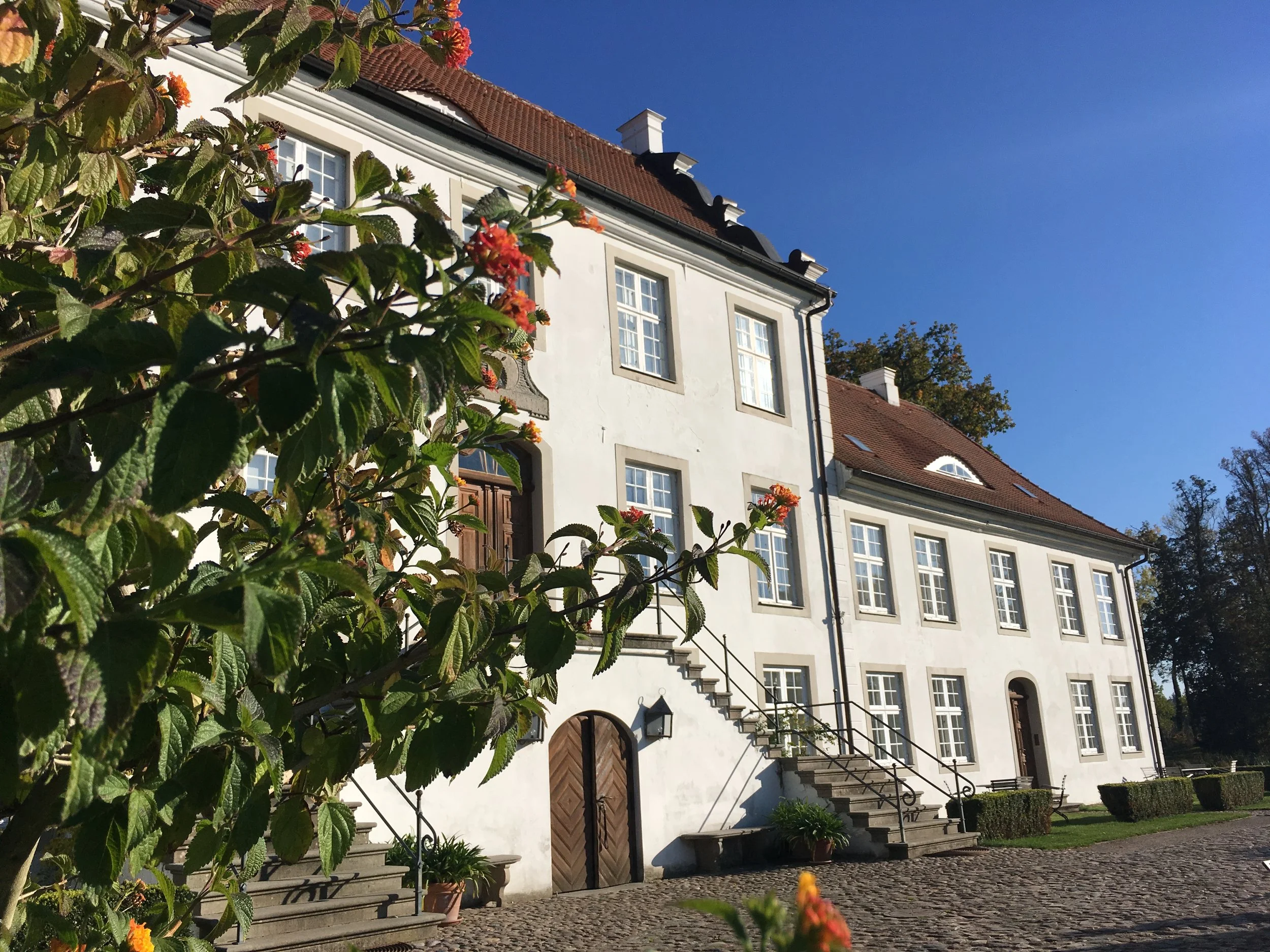 Ein weißes Gebäude mit mehreren Fenstern und rotem Satteldach. Im Vordergrund befinden sich grüne Laubbüsche mit orangefarbenen Blüten. Der Himmel ist klar und blau.