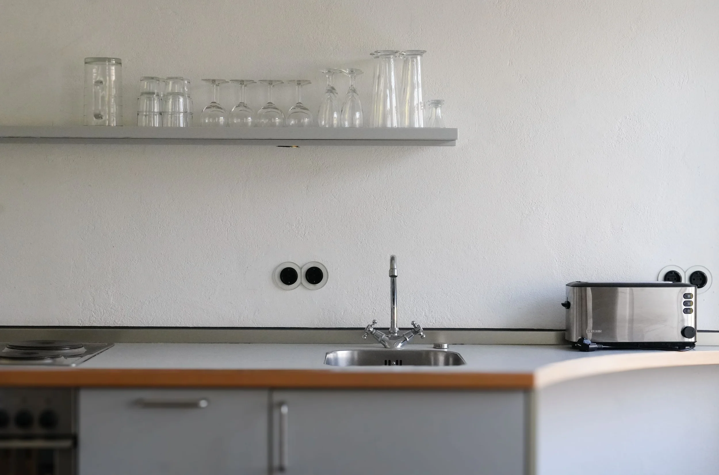 Weiße Küchenzeile mit Spüle, Wasserhahn, einem kleinen Kochfeld, einem Wasserkocher und einem Toaster, an der Wand zwei schwarze Steckdosen, darüber ein Regal mit verschiedenen gläsern.