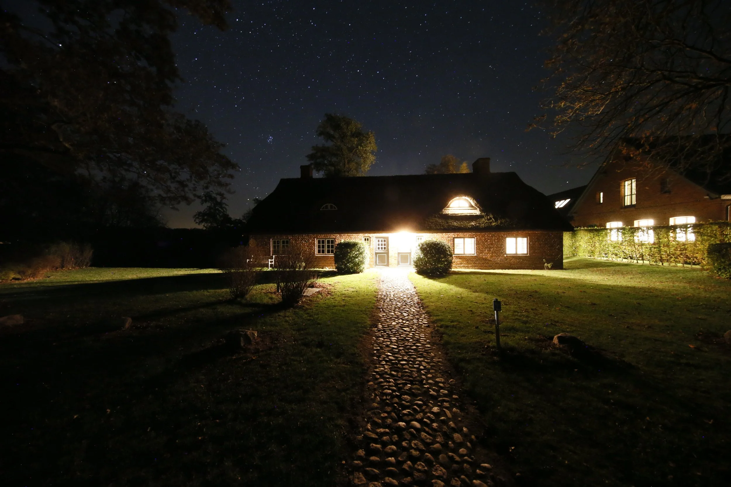 Ein Haus bei Nacht, erleuchtet von innen, mit einem Garten, Sternenhimmel und Sternbilder am Himmel.