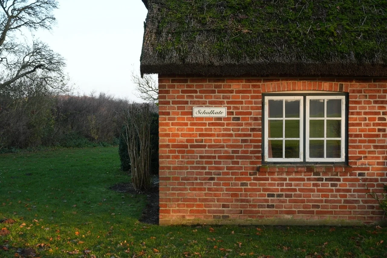 Ein kleines Haus aus roten Backsteinen mit weißem Fenster und einem Schilfdach, das von Bäumen und Wiesen umgeben ist. Ein Schild mit der Aufschrift 'Schullatte' ist an der Hauswand befestigt.