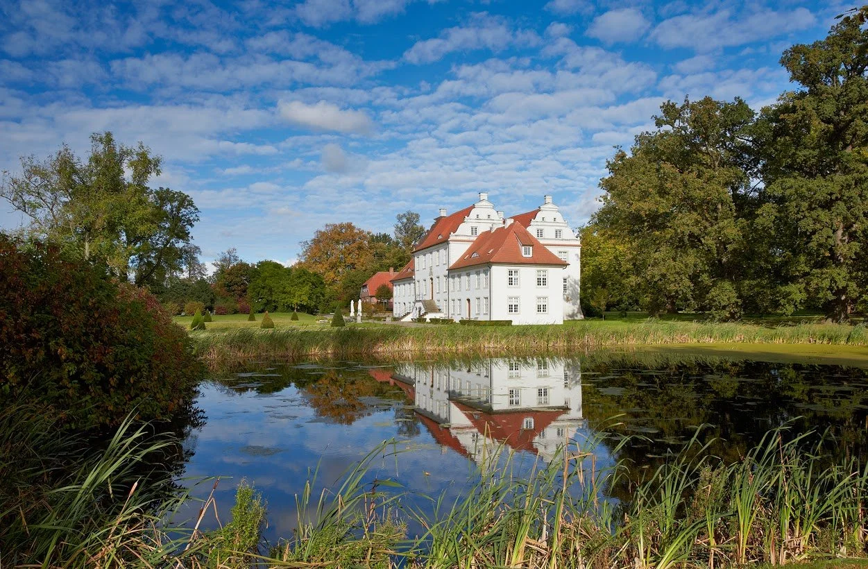 Ein weißes Schloss mit roten Dächern, das von einem See und umgeben von Bäumen in herbstlichen Farben reflektriert wird.