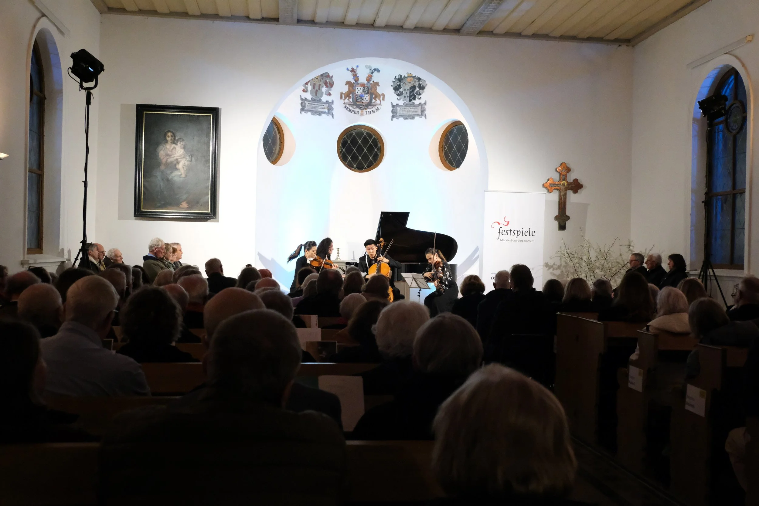Ein Konzert in einer Kirche mit einem Streichquartett auf der Bühne, beobachtet von einem Publikum, im Hintergrund religiöse Kunst und Wappen an der Wand.