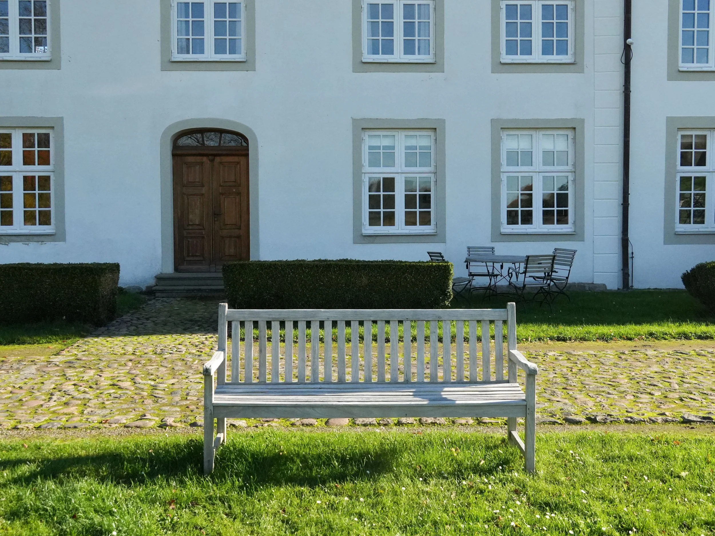 Ein weißes Gebäude mit mehreren Fenstern, ein Holzport mit einer Tür, ein gepflasterter Weg, eine Bank, ein kleiner Zaun und einige Büsche im Vordergrund.
