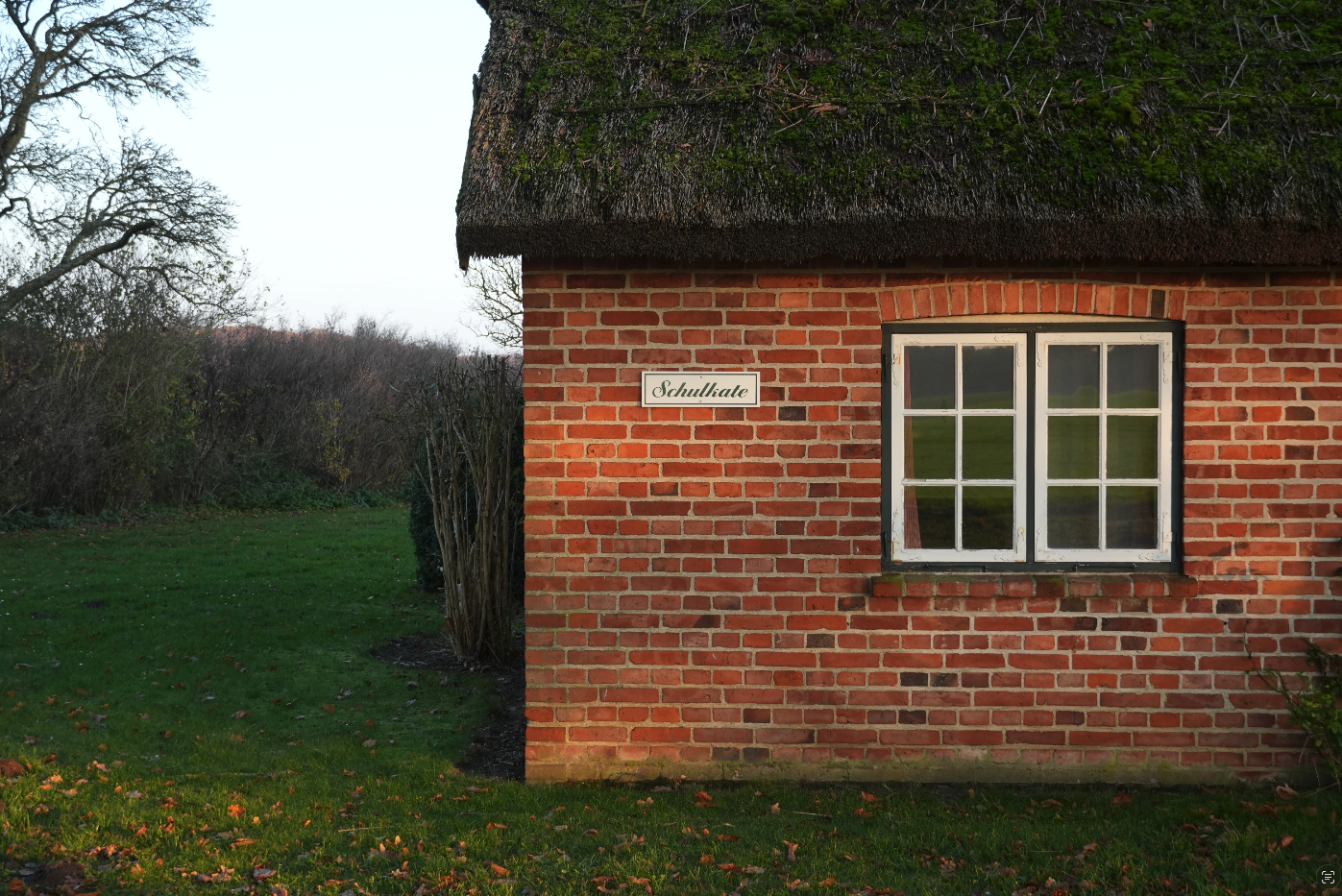 Ein kleines, rotes Backsteinhaus mit weißem Fenster und Schilfdach, umgeben von grünem Rasen und Büschen, mit einem Straßenschild 'Schullate' auf der Hauswand.