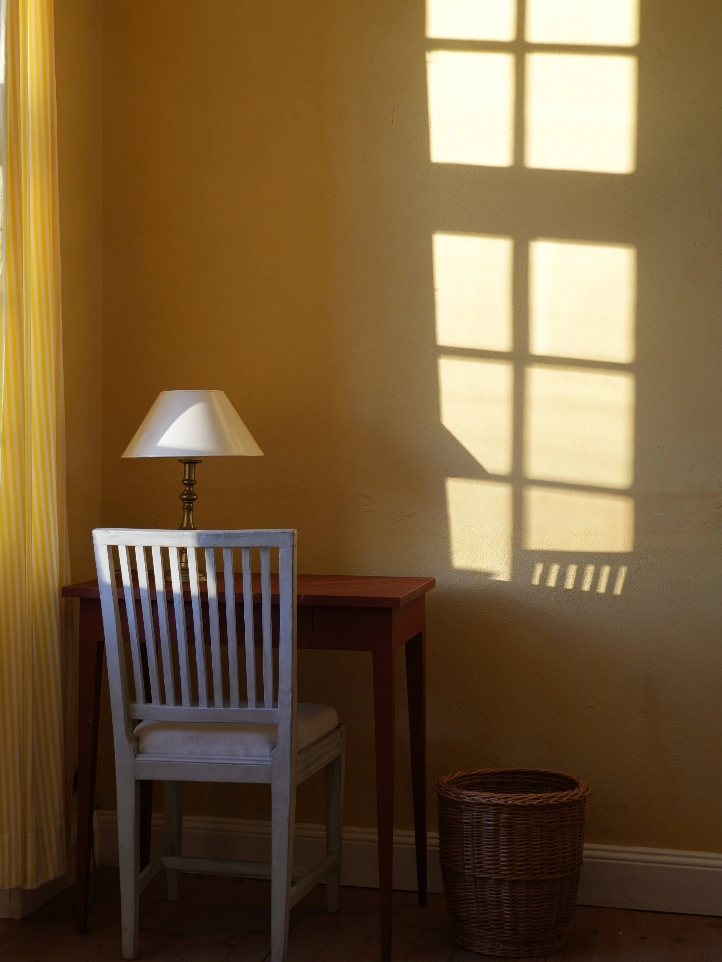 Lampenlicht wirft Schatten eines Fensters auf die gelbe Wand. Im Raum steht ein runder Holztisch mit einem weißen Stuhl und einem Korb daneben.