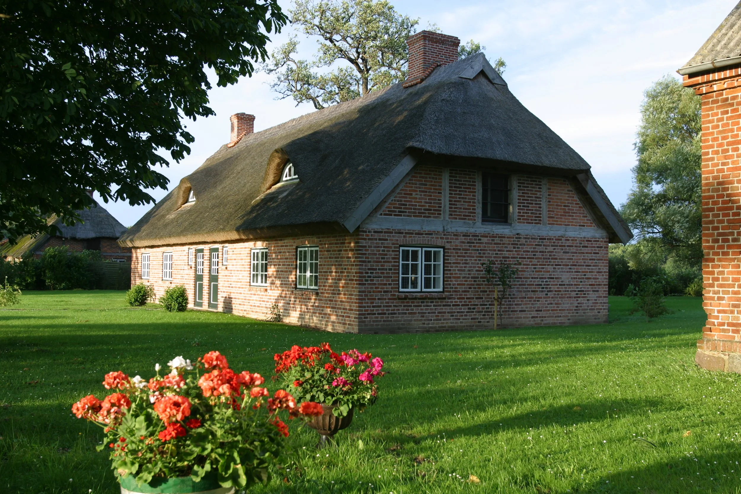 Ein traditionelles rotes Backsteinhaus mit Strohdach, umgeben von einem grünen Garten mit bunten Blumen. Es steht im Freien bei sonnigem Wetter, mit einem Blattbaum auf der linken Seite und einem weiteren Backsteingebäude an der rechten Bildseite.