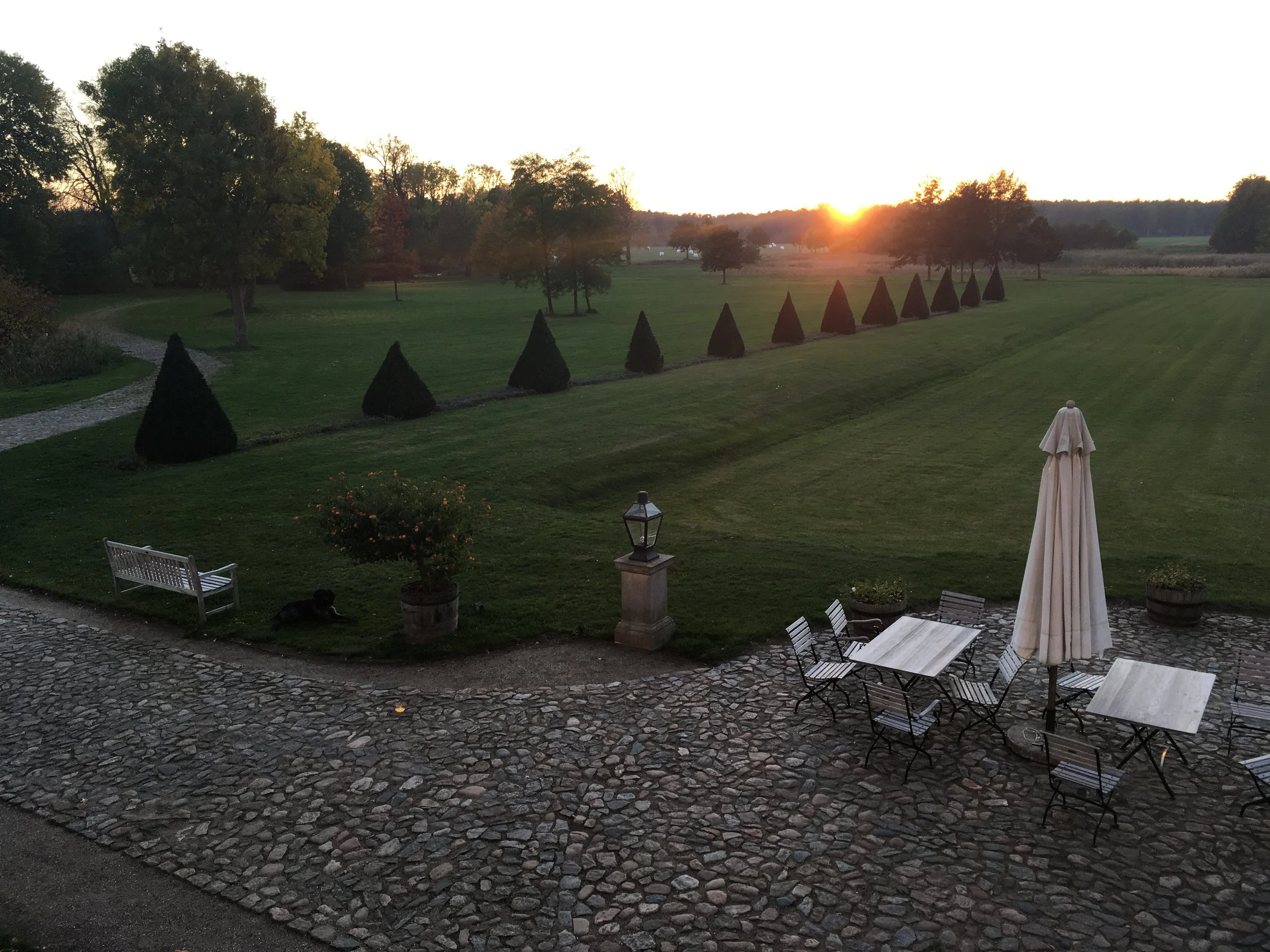 Garten im Sonnenuntergang mit Rasen, Bäumen, einem weißen Sonnenschirm, Sitzgelegenheiten und einem Weg aus Kies.