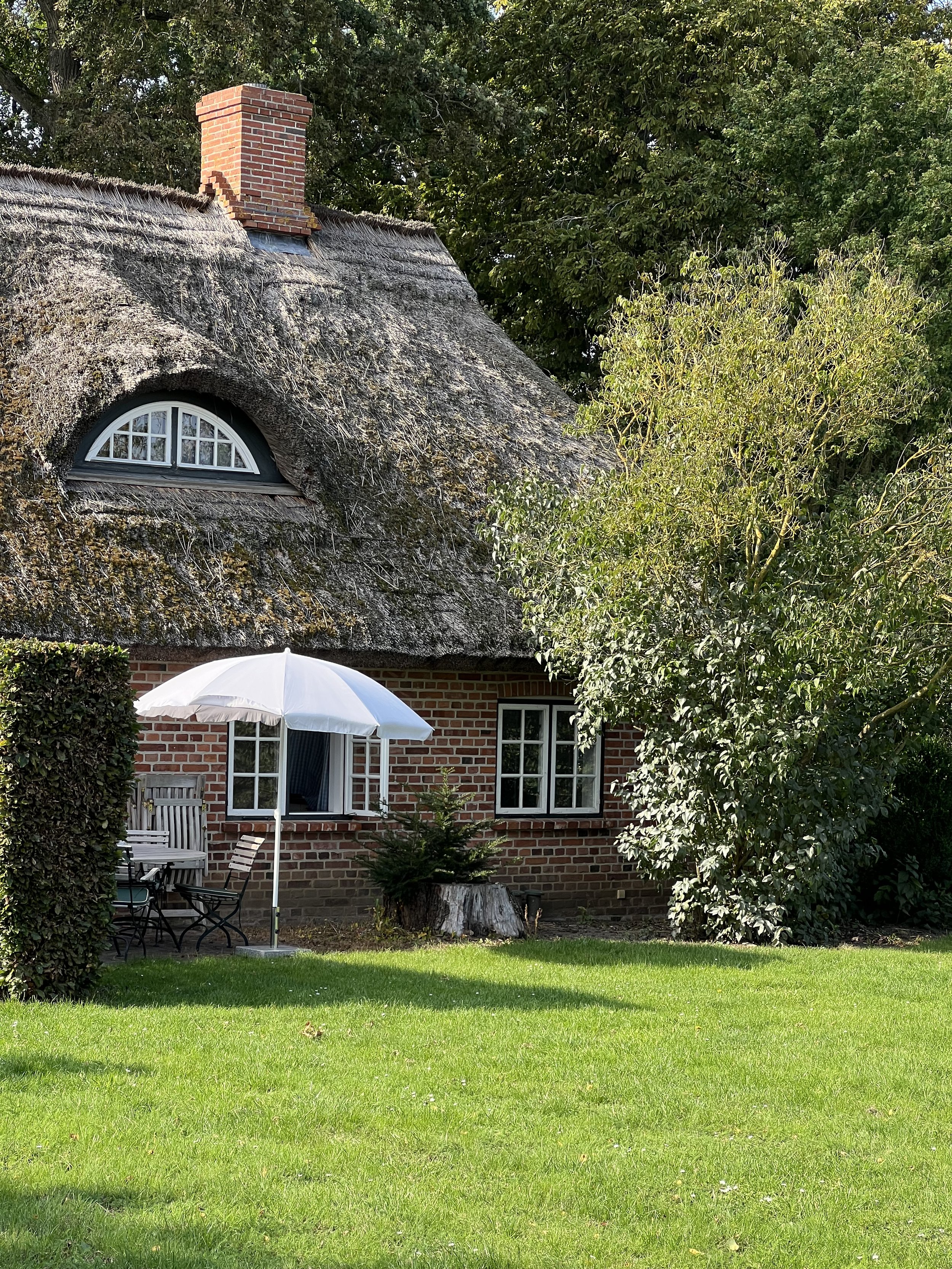 Ein Haus mit einem Schilfdach, Balkon und weißen Fensterläden, im Garten stehen eine weiße Sonnenbank und ein Schirm, umgeben von Bäumen und grüner Wiese.