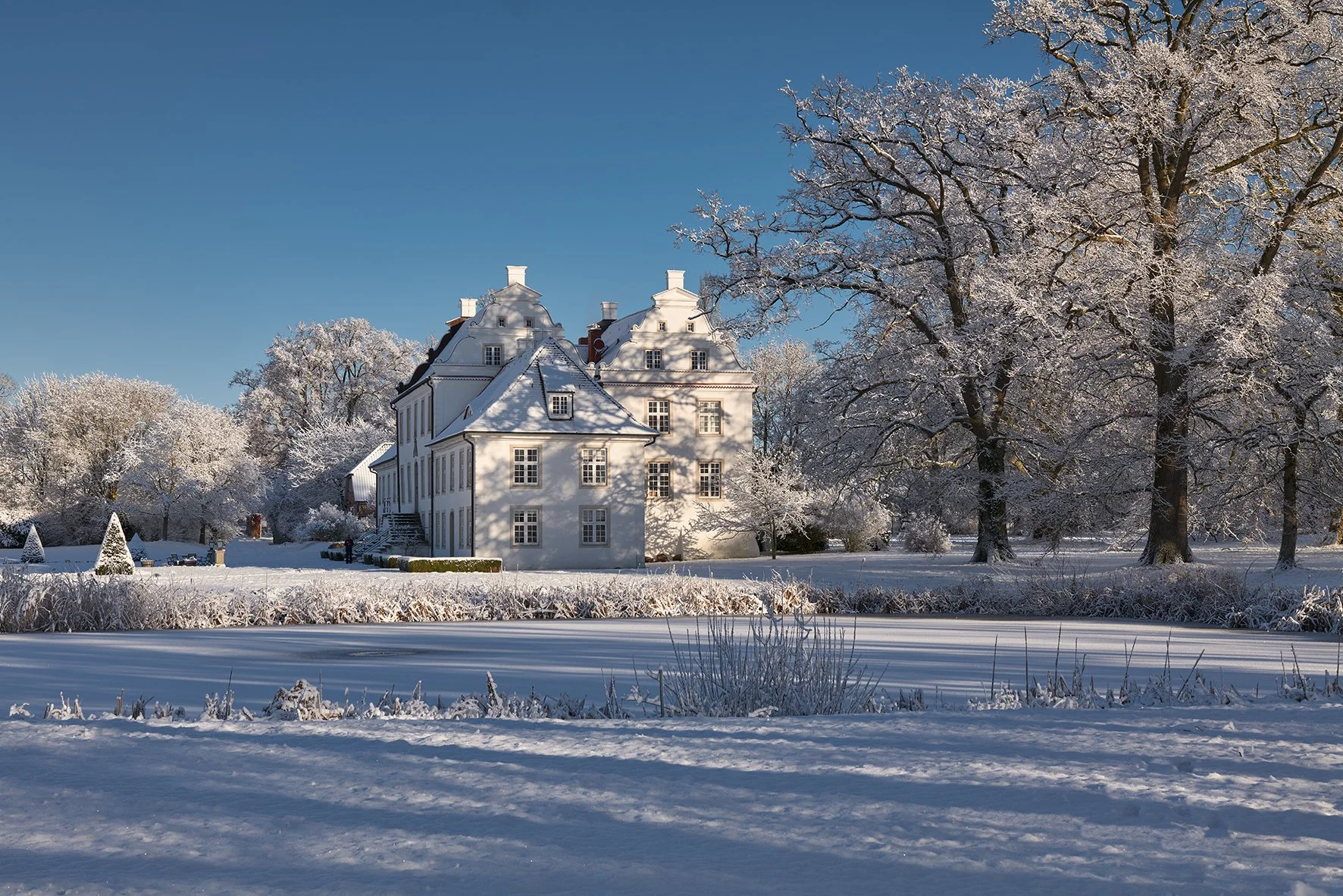 Ein weißes Schloss im verschneiten Park, umgeben von frostbedeckten Bäumen und einem zugefrorenen Teich bei klarem blauen Himmel.