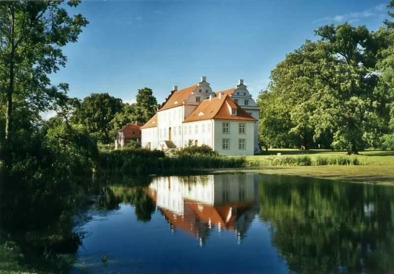 Ein großes weißes Schloss mit roten Dächern liegt an einem See, umgeben von grünen Bäumen bei sonnigem Wetter.
