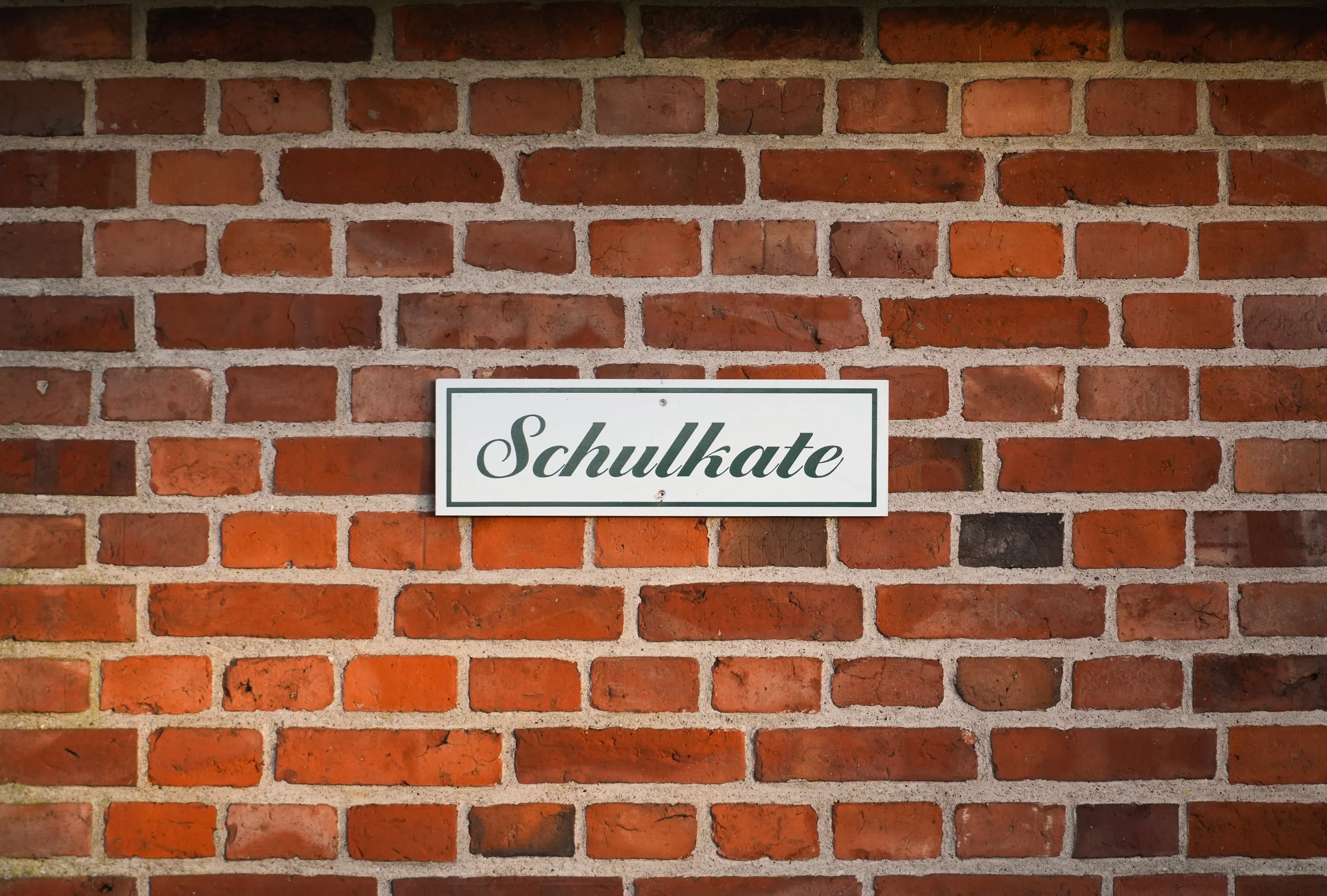 Ein roter Backsteinwand mit einem weißen Schild, auf dem das Wort "Schulkate" steht.