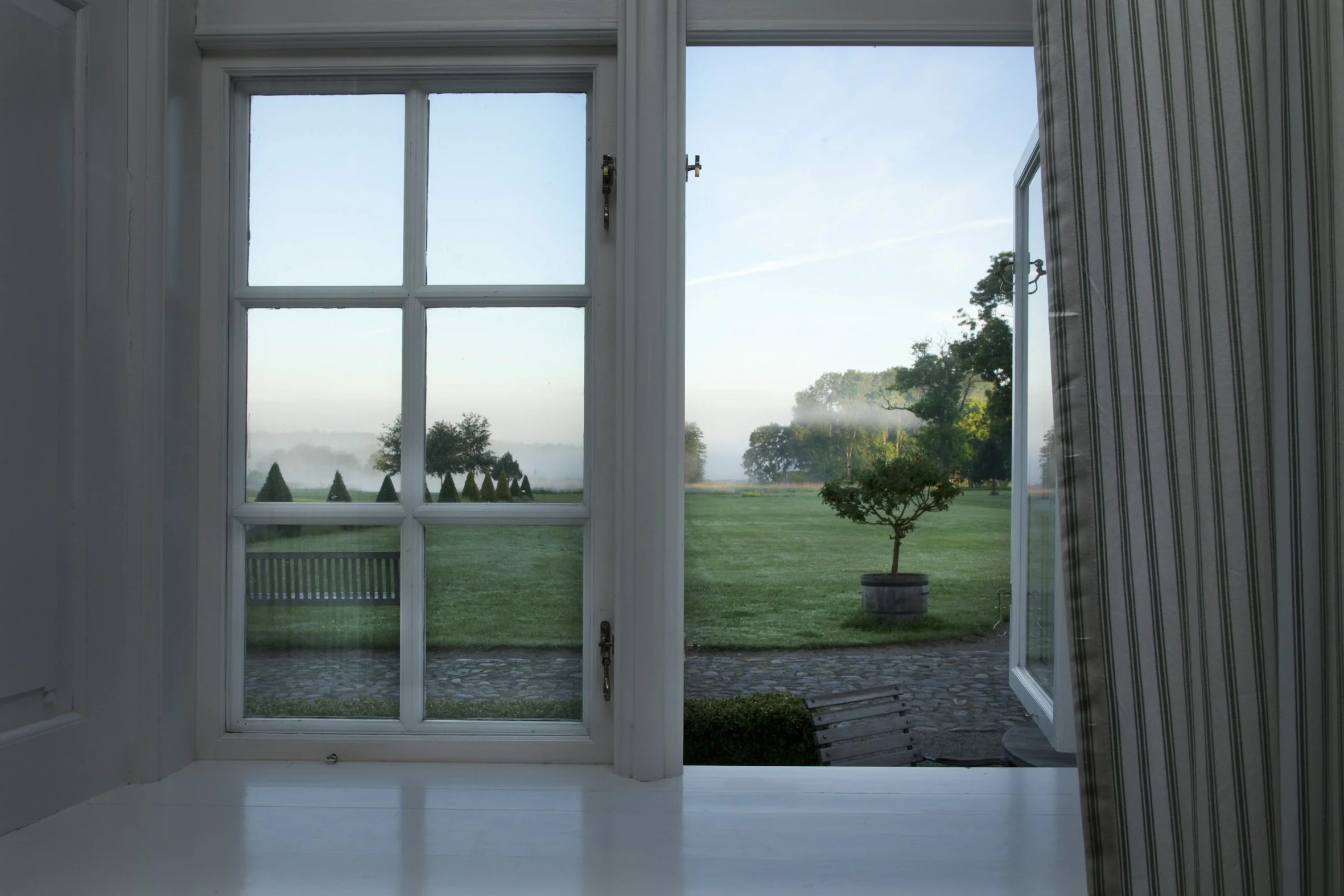 Blick aus einem offenen Fenster auf eine grüne Wiese mit Bäumen im Hintergrund an einem clearen Tag.