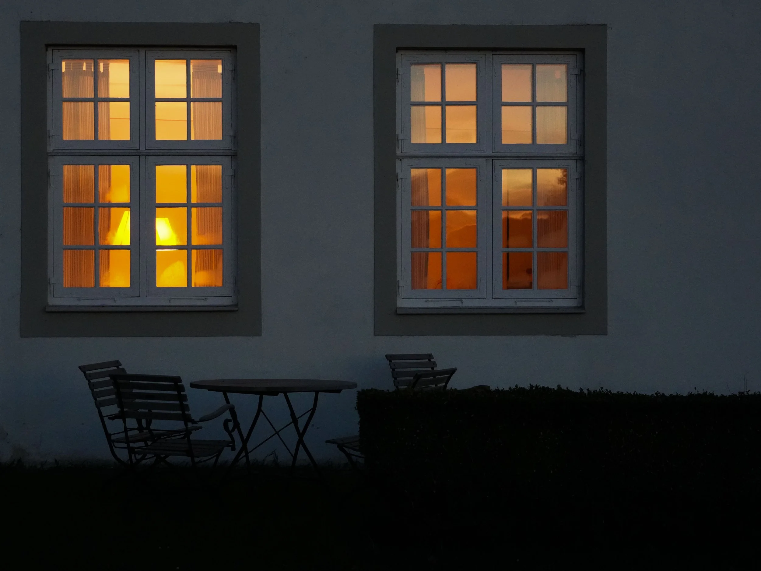 Blick auf zwei beleuchtete Fenster eines Gebäudes bei Dämmerung, außen davor ein Tisch und Stühle im Garten.