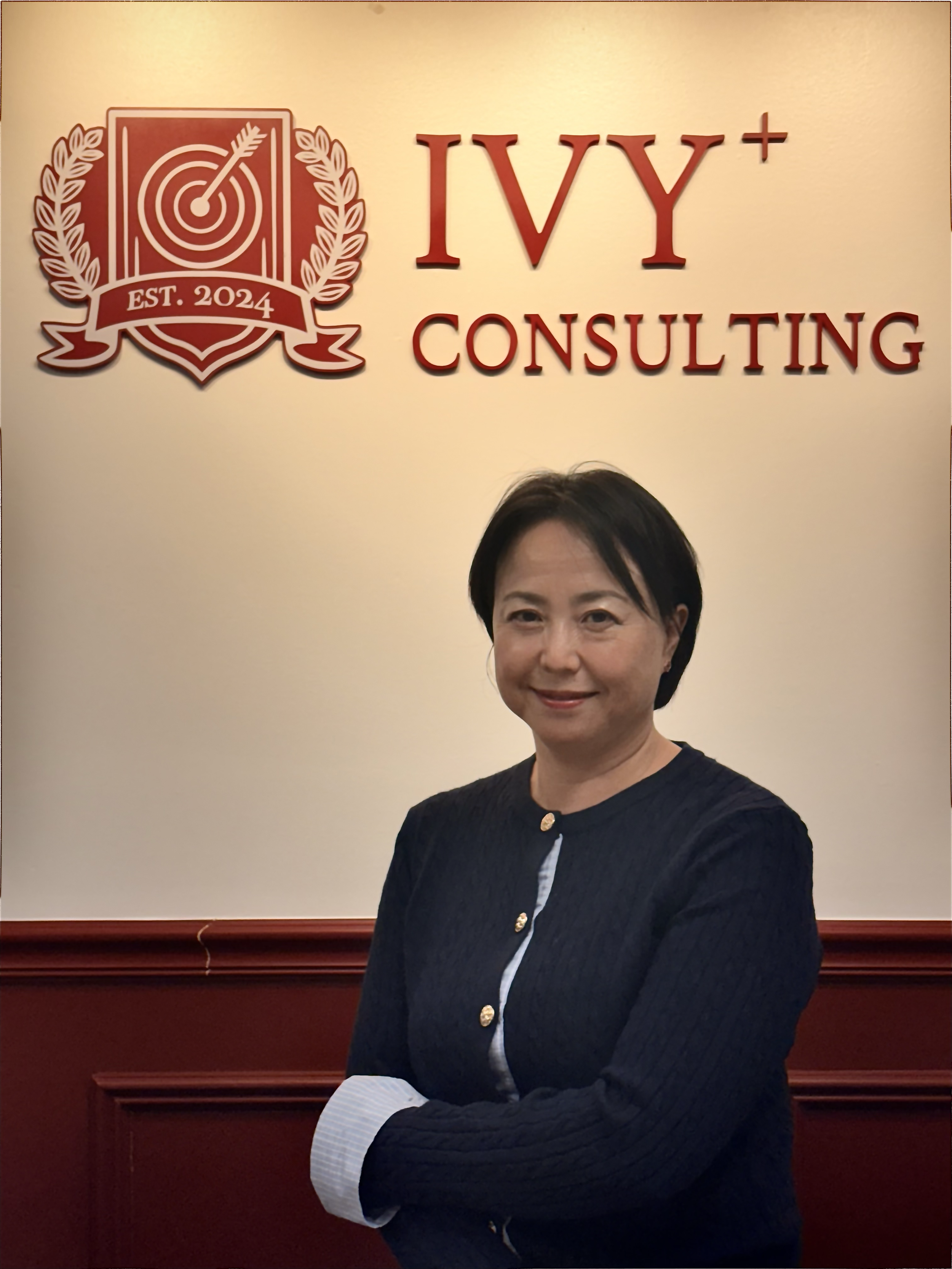 A woman standing confidently in front of a wall with a logo and the words 'IVY+ Consulting.' The logo includes a target with an arrow, a shield, and laurel wreaths, with 'EST. 2024' written inside. The woman is wearing a dark jacket with gold buttons and has her arms crossed.