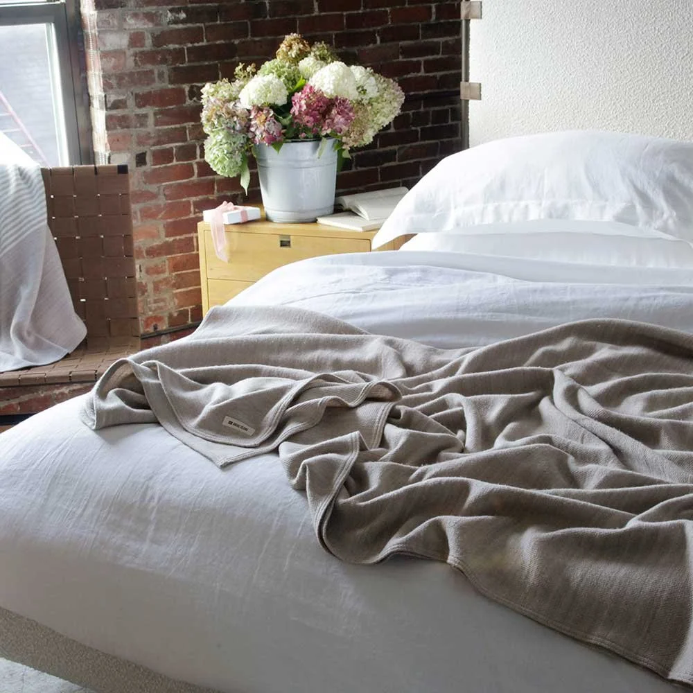 Unmade bed with white sheets and pillows, a beige blanket draped over it, a wooden bedside table with a large floral arrangement in a white pot, an open book, and a pink ribbon. There is a brick wall and a window in the background.