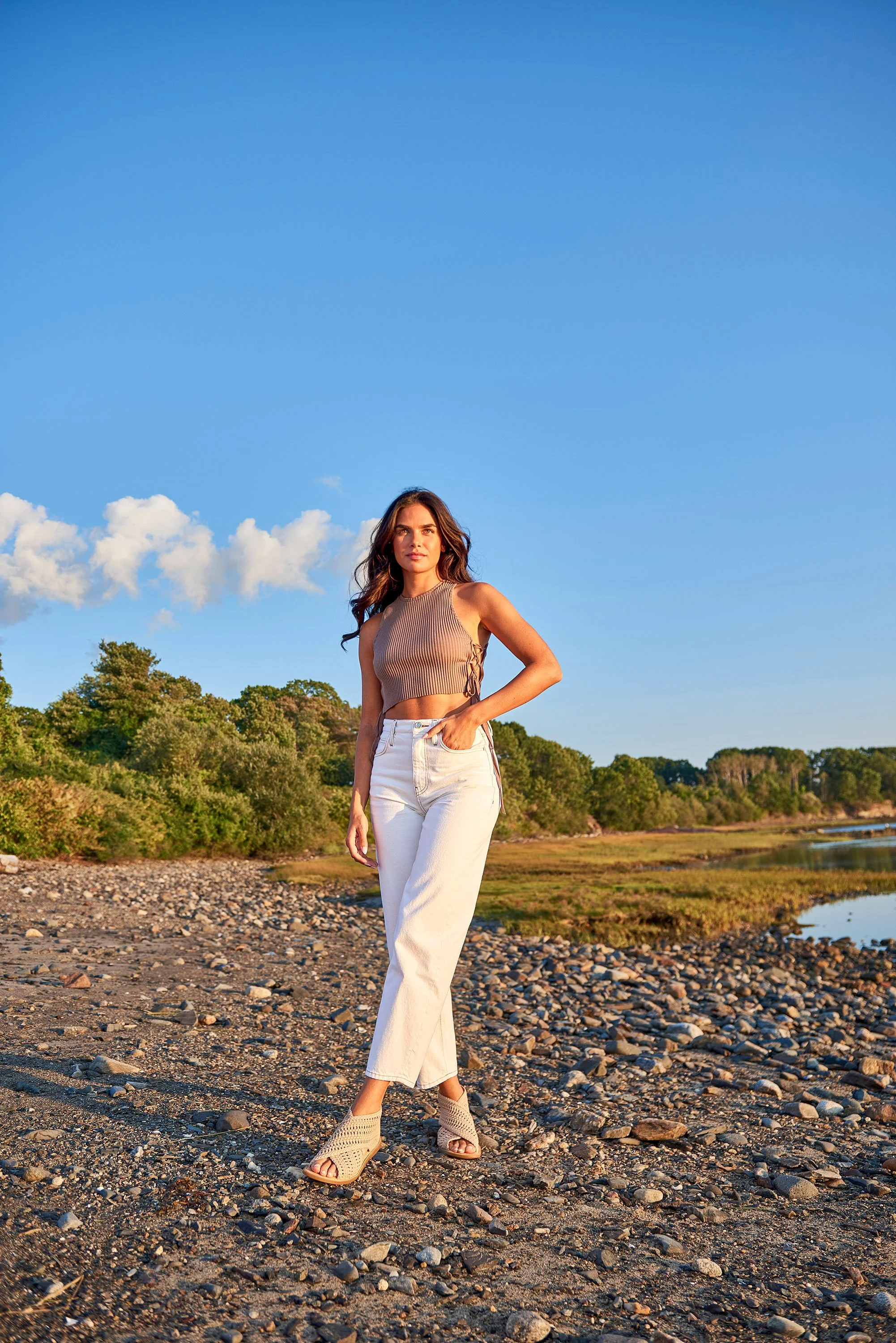 A woman in a beige sleeveless crop top and white pants standing on a rocky beach during sunset, with trees and water in the background.