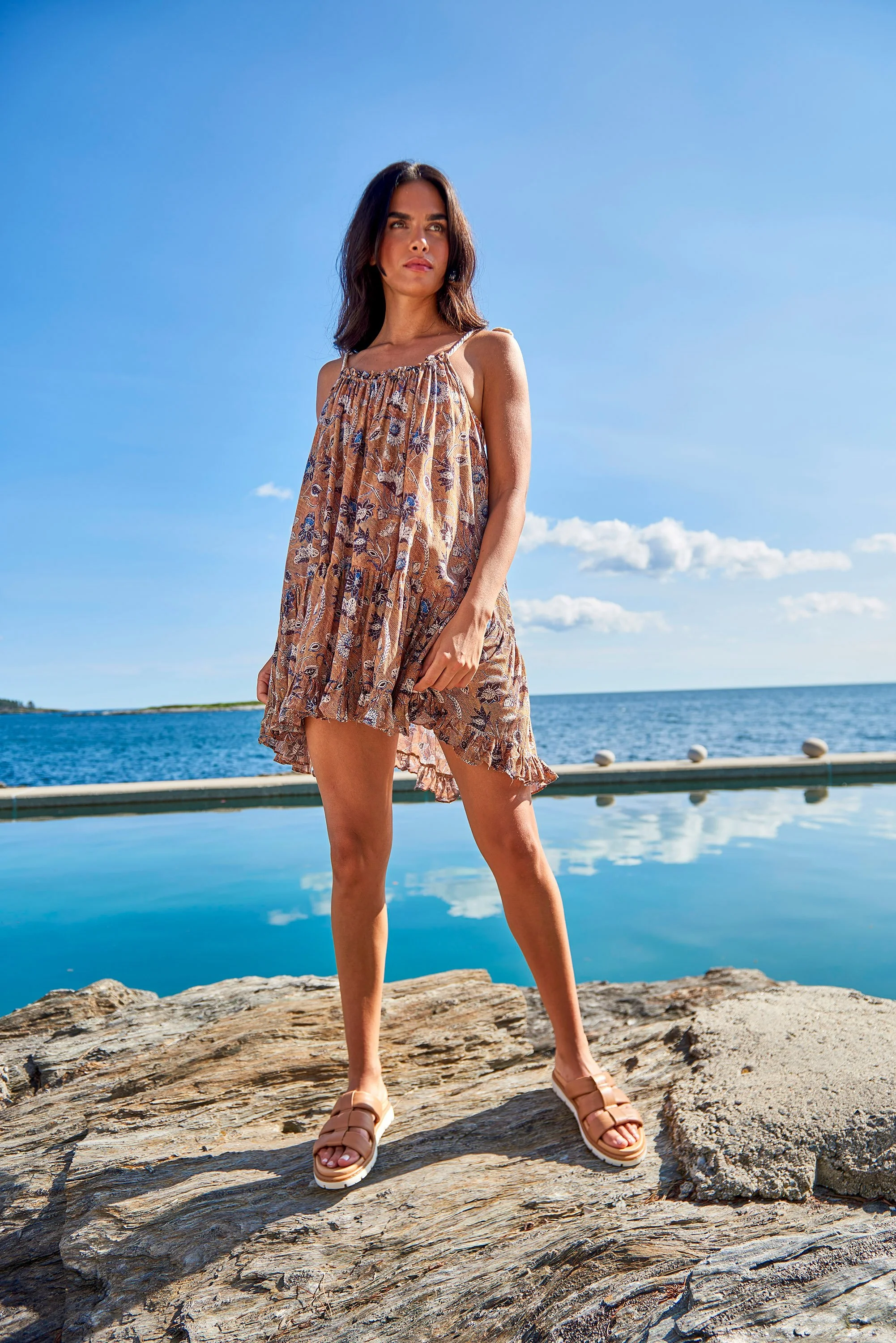 Woman standing on rocks near water on a sunny day, wearing a floral dress and sandals, with ocean and blue sky in the background.