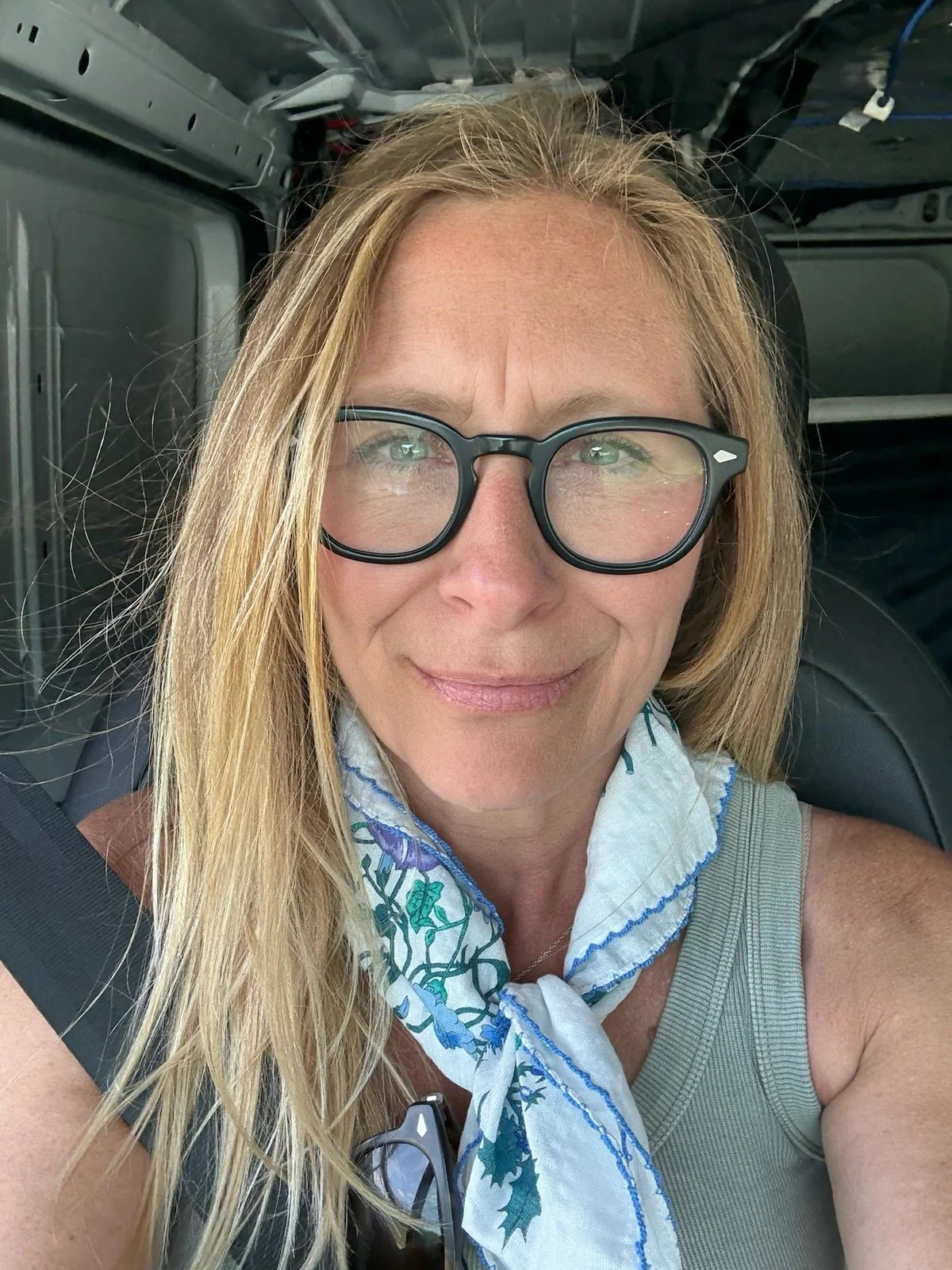 A woman with blonde hair, glasses, and a blue and white scarf, sitting in a vehicle with a seatbelt on.