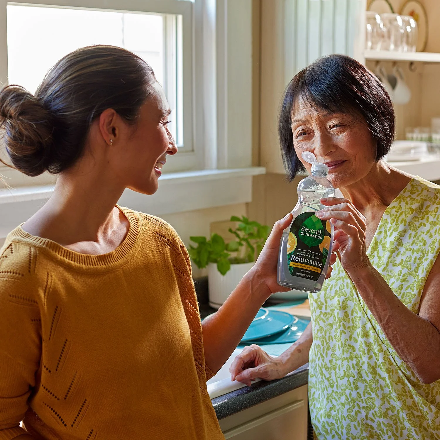 An older woman is smelling a bottle of Seventh Generation Rejuvenate hand soap held by a younger woman in a kitchen with natural light and green plants.