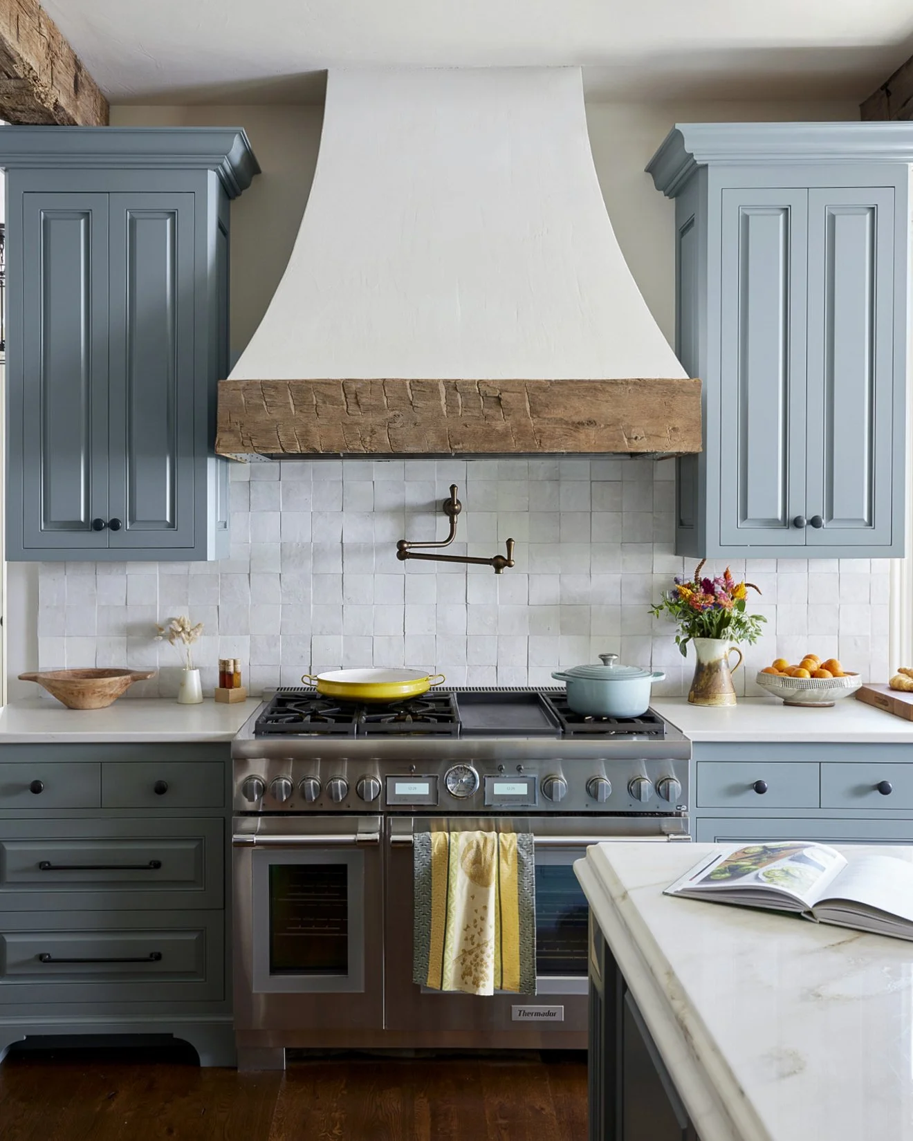 Kitchen with gray cabinets, a stainless steel range, a yellow cast-iron skillet, a light blue pot, a vase with colorful flowers, and open cookbook on marble countertop.