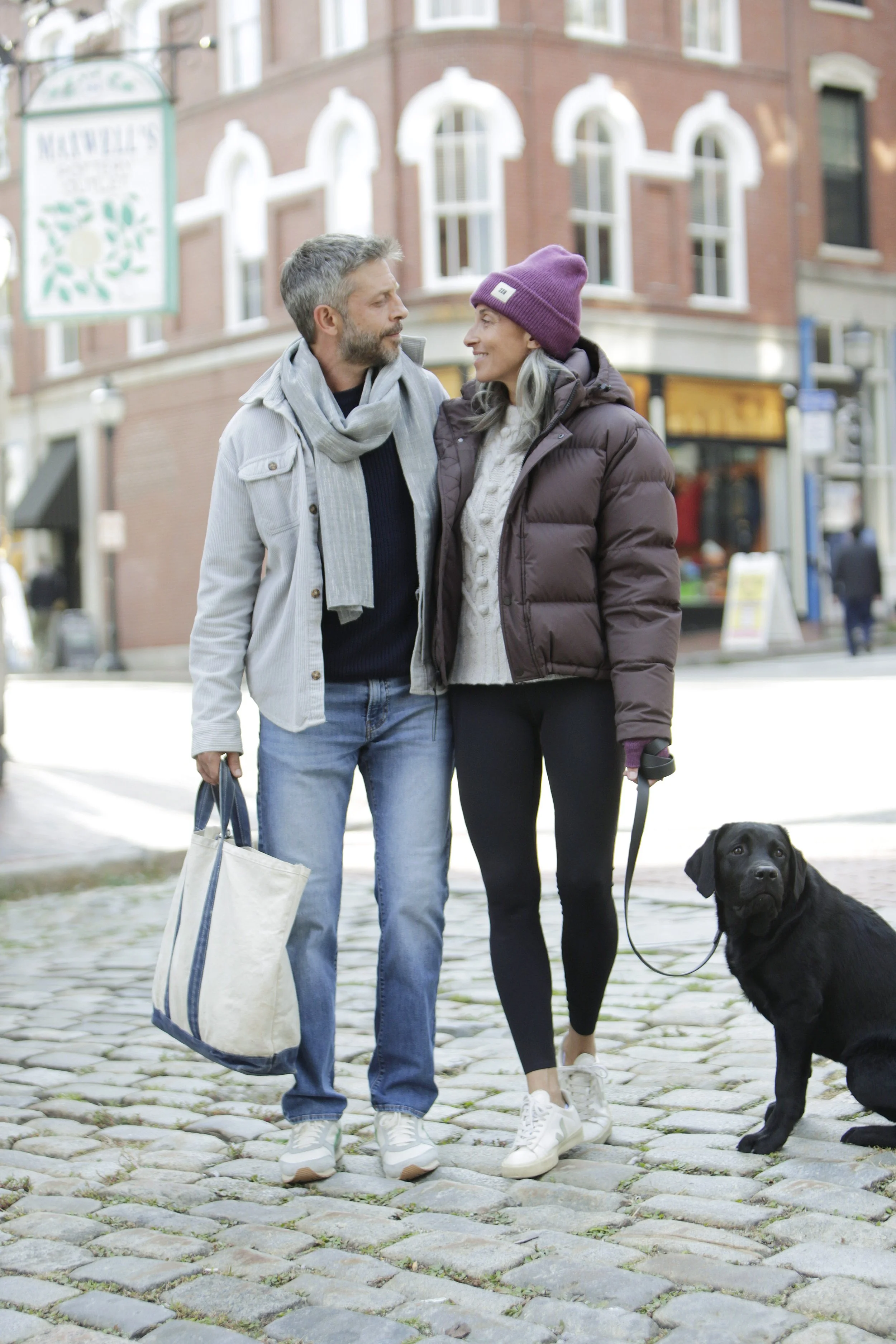 A man and woman walking together on a cobblestone street with a black dog on a leash, talking and smiling, in a city scene with brick buildings in the background.