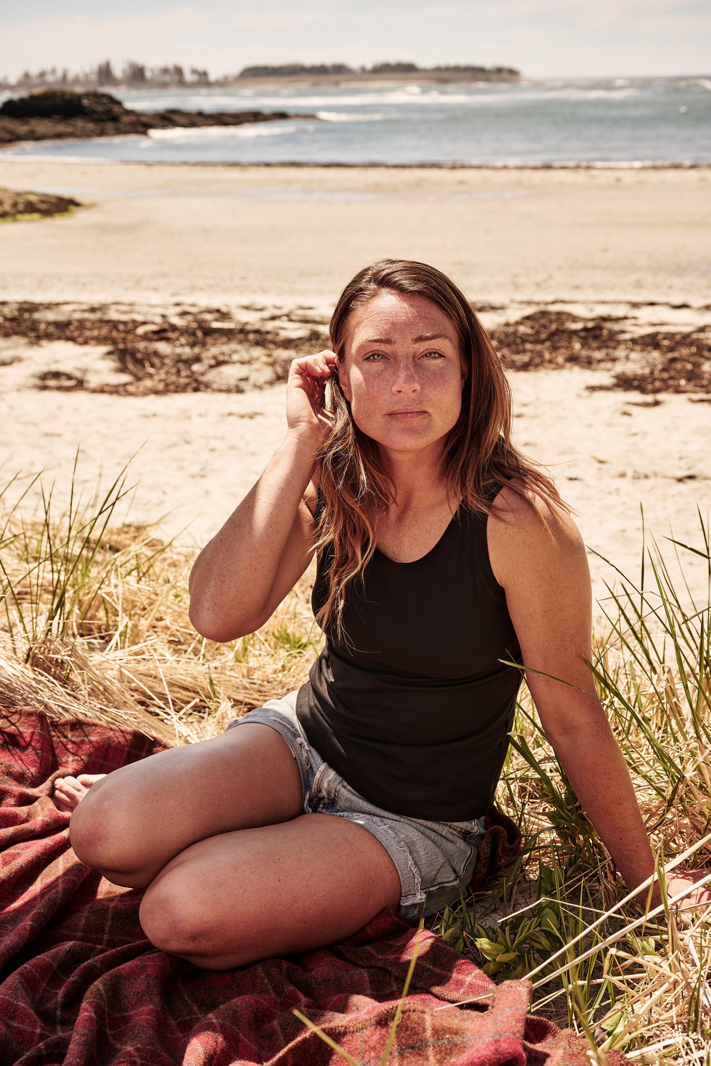 A woman sitting on a blanket at the beach, with sand, ocean waves, and a distant shoreline in the background.