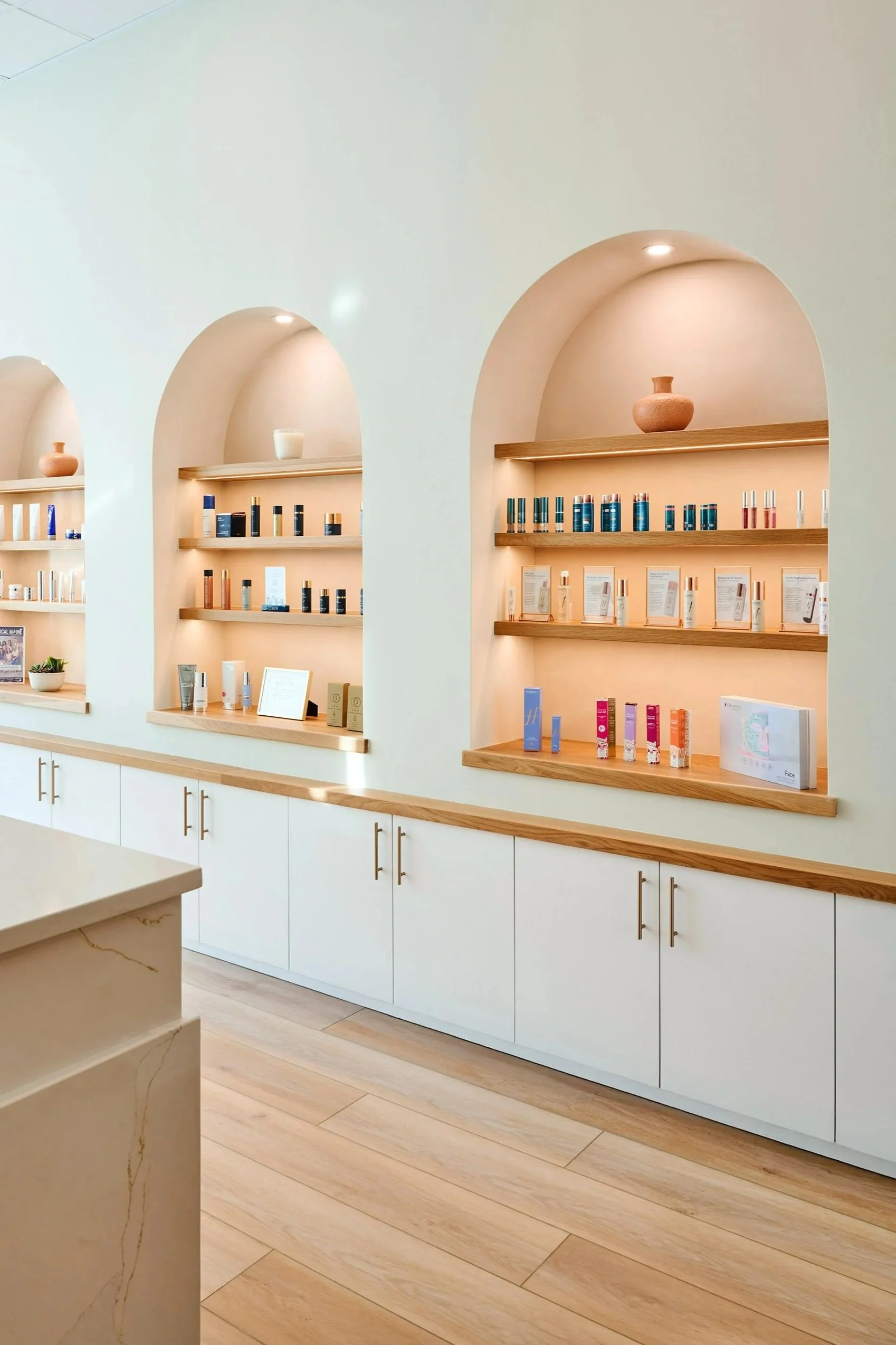 Interior of a retail beauty or skincare store with display shelves built into arched wall niches, featuring various skincare products and decorative pottery, with white cabinets and light wood flooring.
