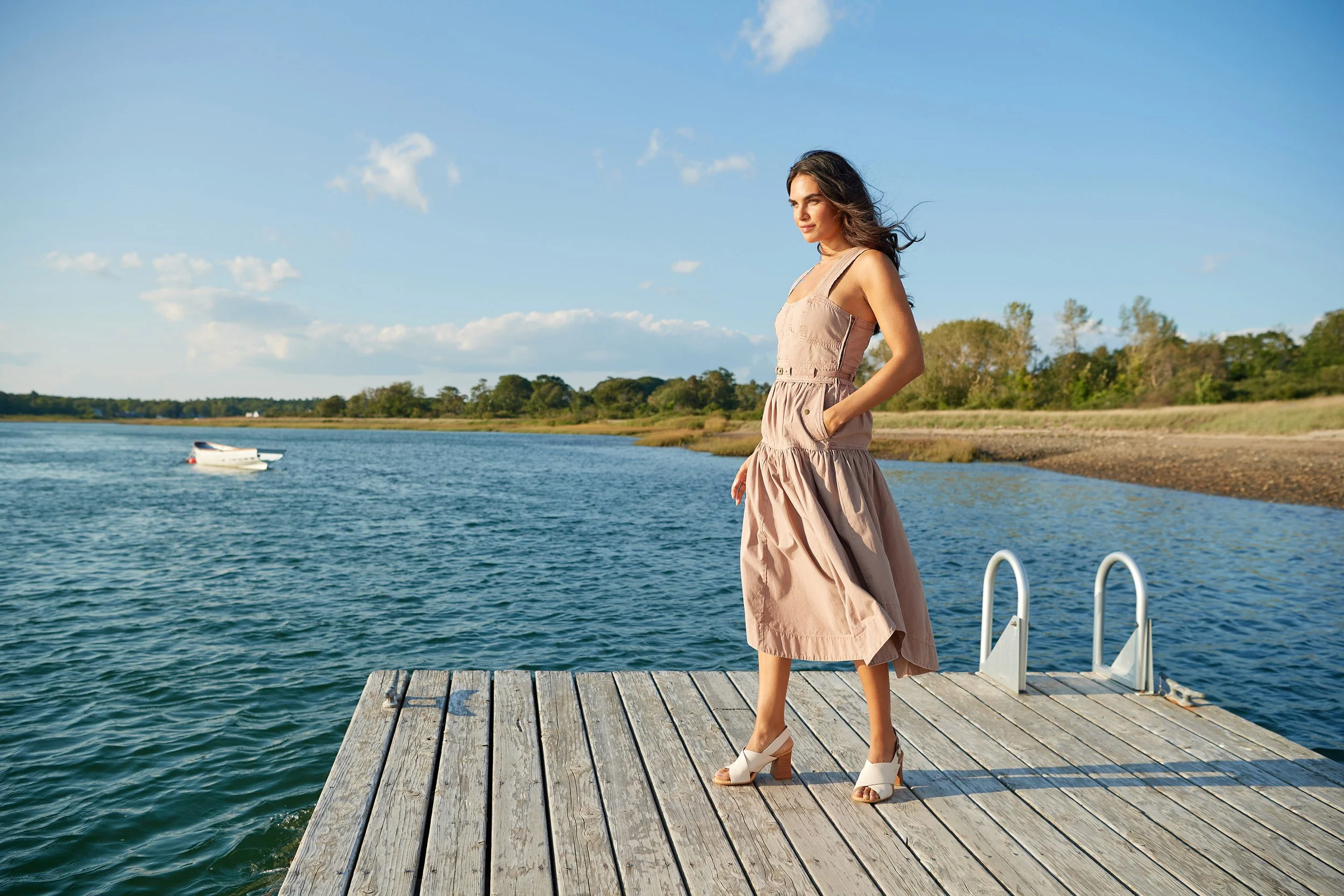 A woman in a beige dress and white high heels standing on a wooden dock by a body of water, with trees and a boat in the background on a sunny day.