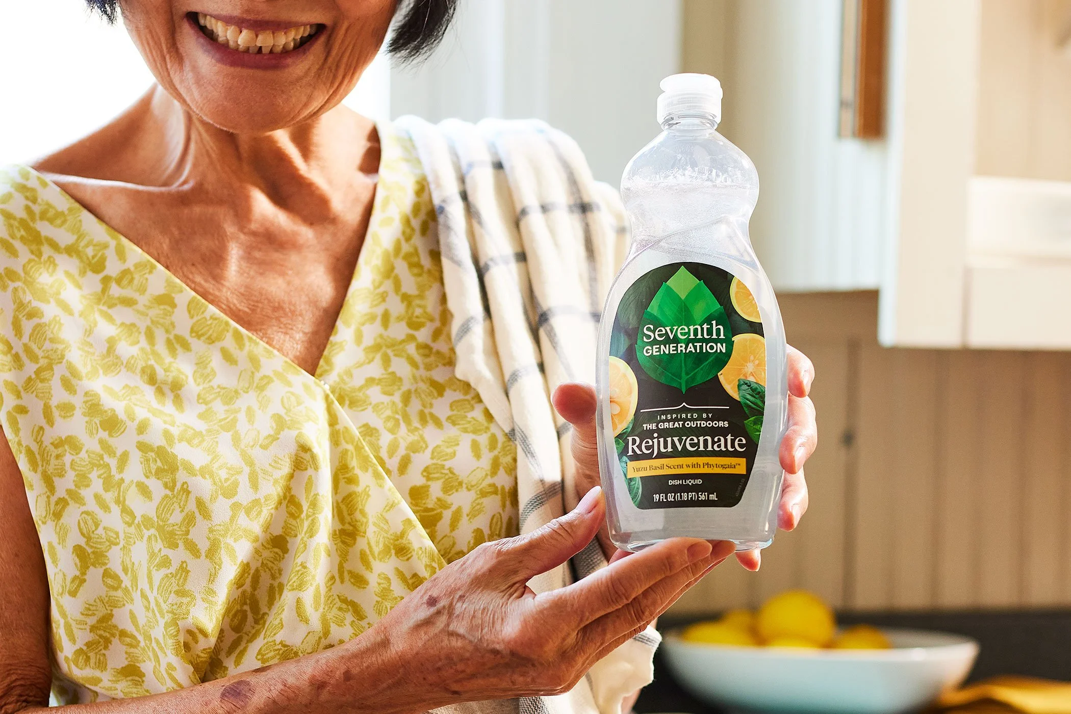 A woman holding a bottle of Seventh Generation Rejuvenate dish liquid in a kitchen.